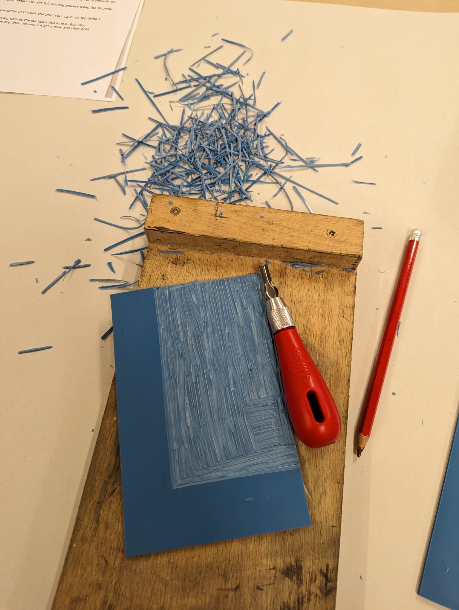 Photo of a carved lino piece in the shape of an 'L', situated next to a cutting tool and pencil.