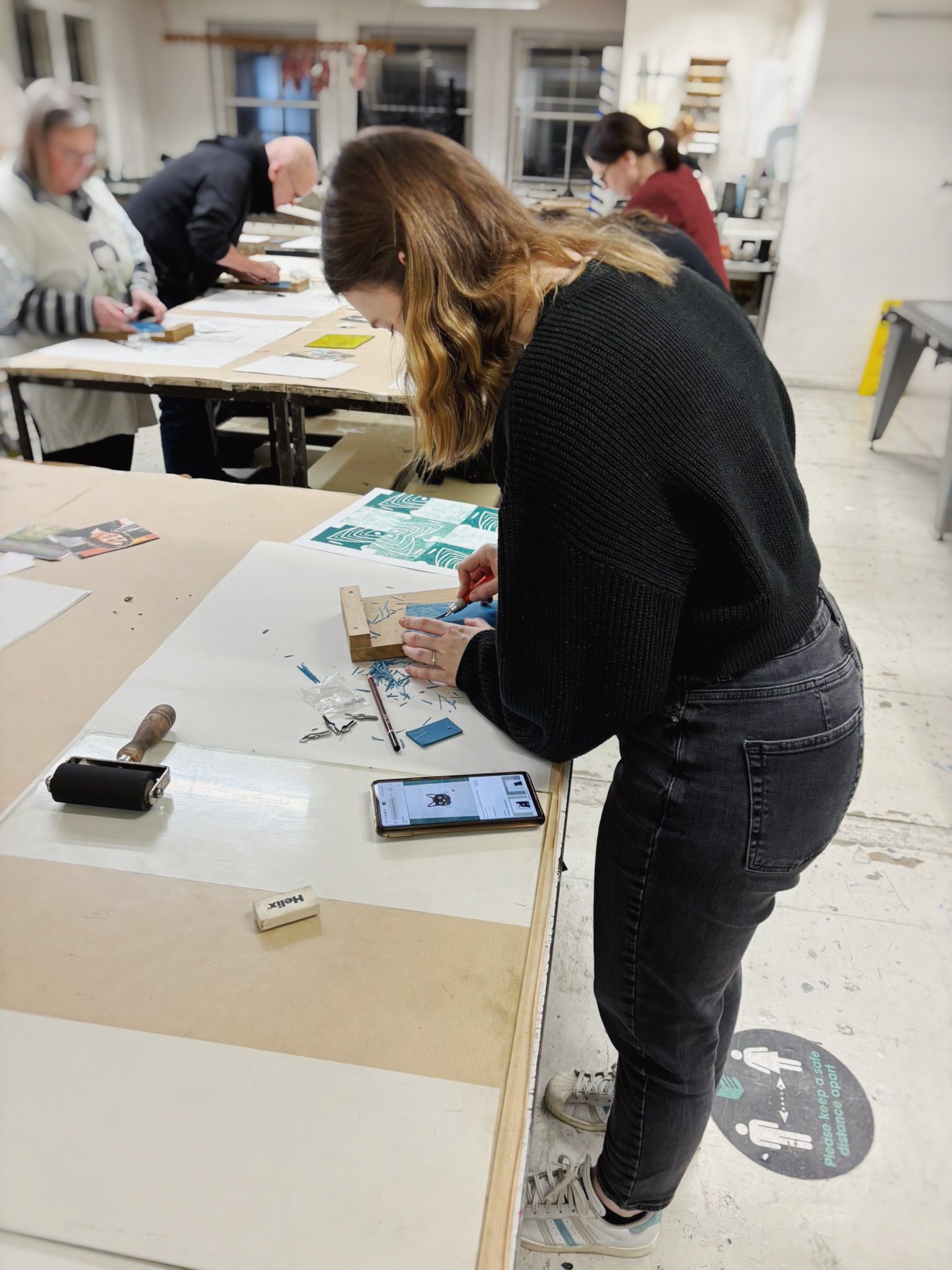 Photograph of a woman standing in classroom in front of a table, leaning over and cutting lino with a lino cutting tool. People can be seen in the background but are out of focus.