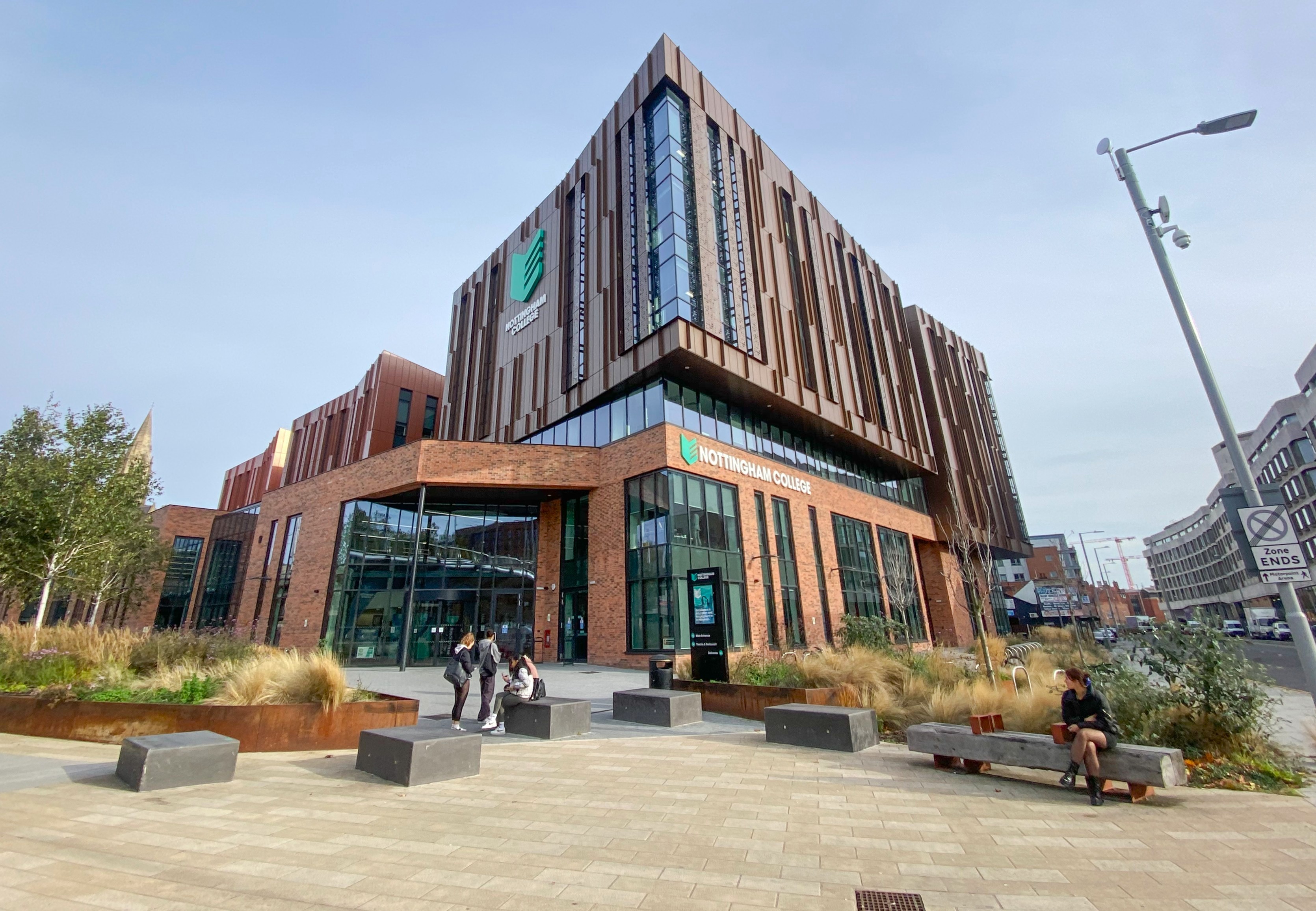 Photograph of a large building with an orange toned exterior against a blue sky, surrounded by plants.