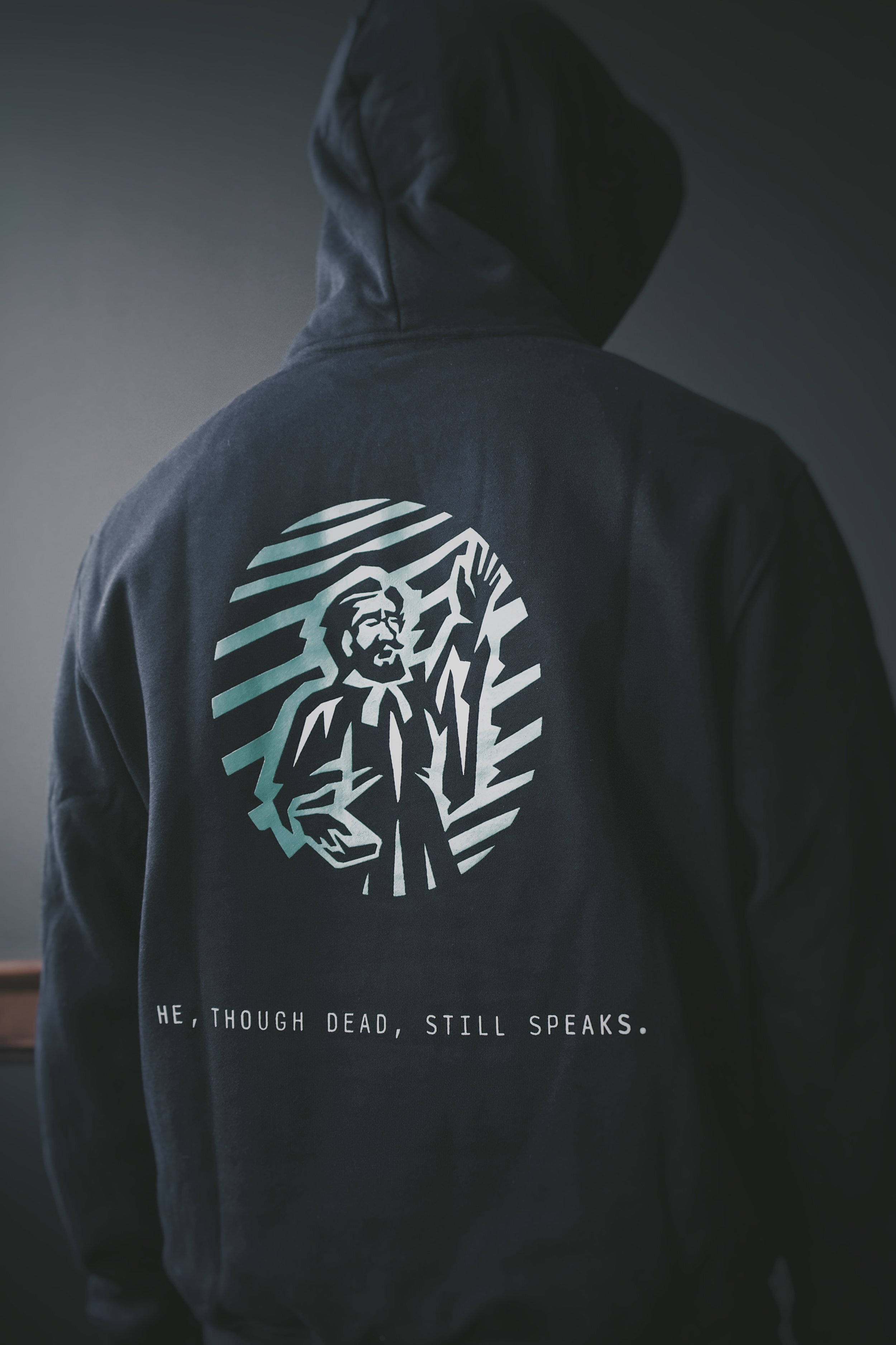 Back of a black hoodie featuring a stylized preacher illustration and the text ’He, though dead, still speaks.’