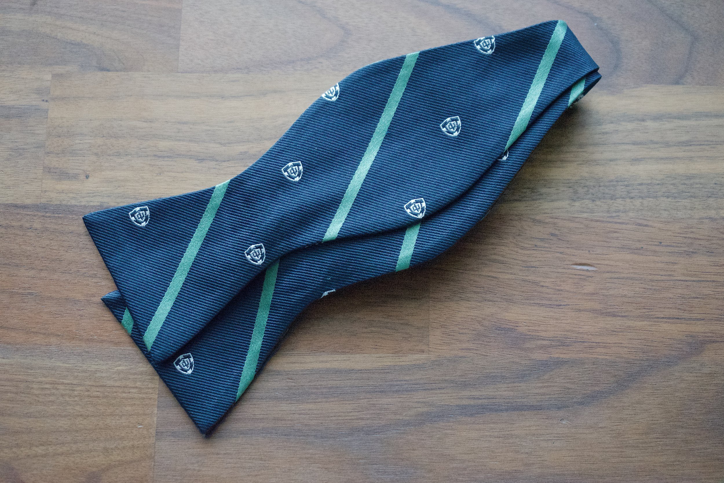 Navy bow tie with green diagonal stripes and small crest emblems laid flat on a wooden surface.