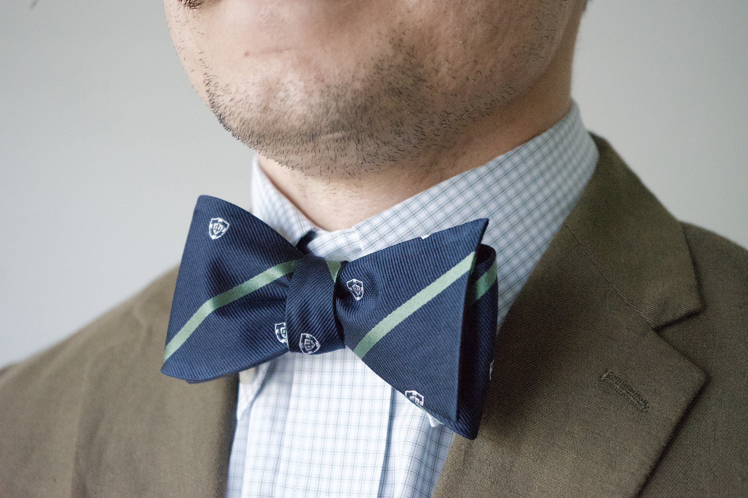 Navy bow tie with green diagonal stripes and crest pattern, worn with a checkered dress shirt and blazer.