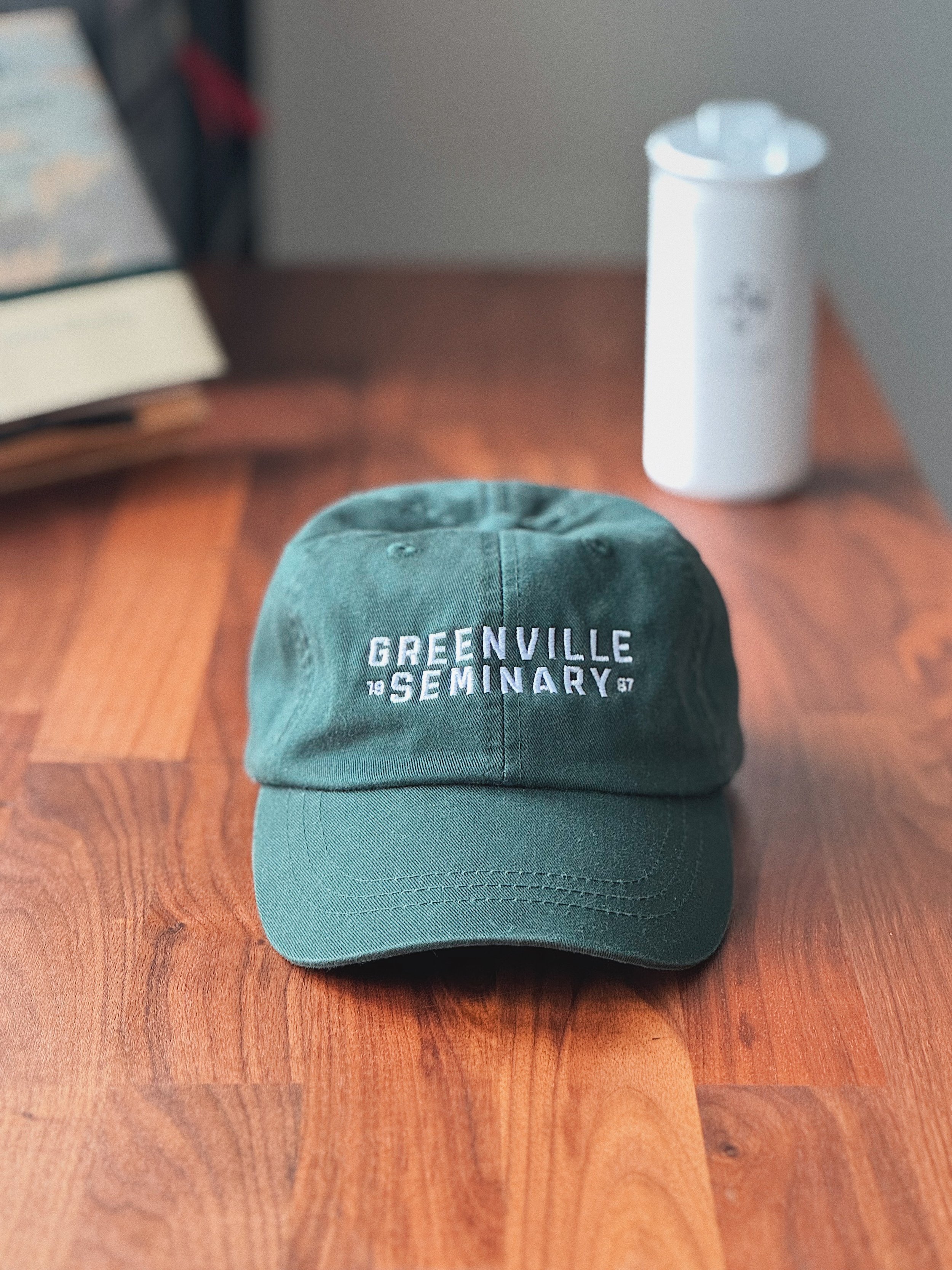 Green Greenville Seminary baseball cap with embroidered text displayed on a wooden surface.