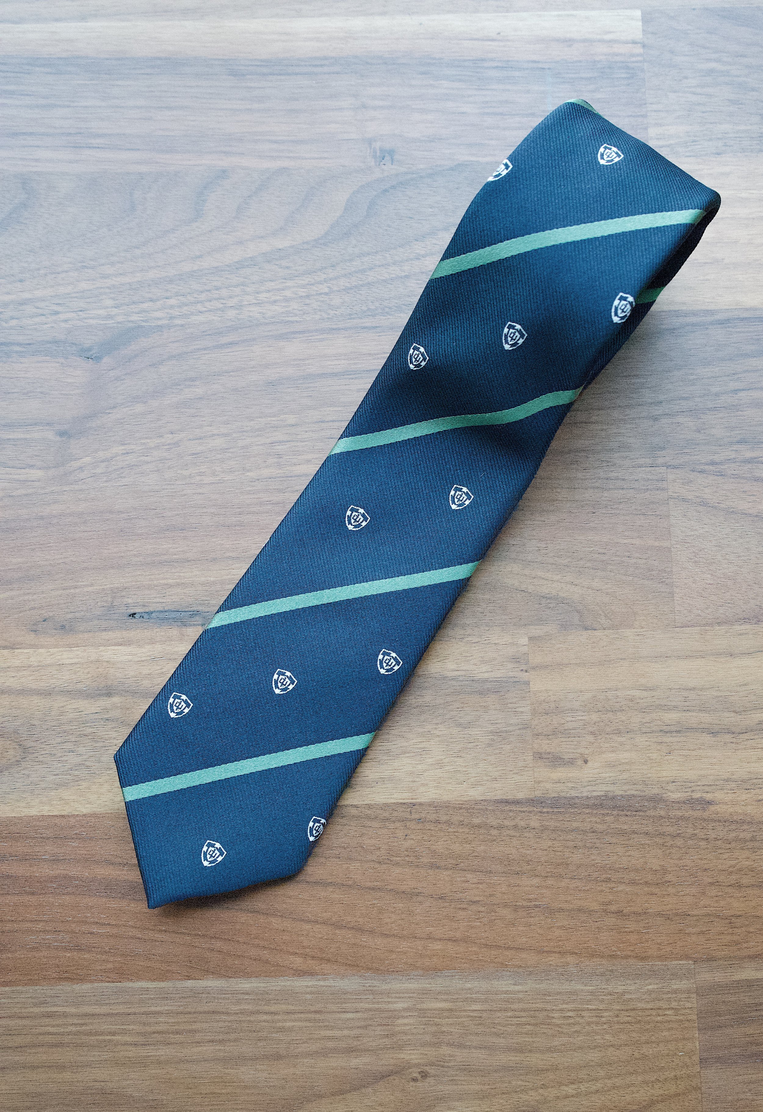 Angled view of a navy necktie with green diagonal stripes and crest emblems on a wooden surface.