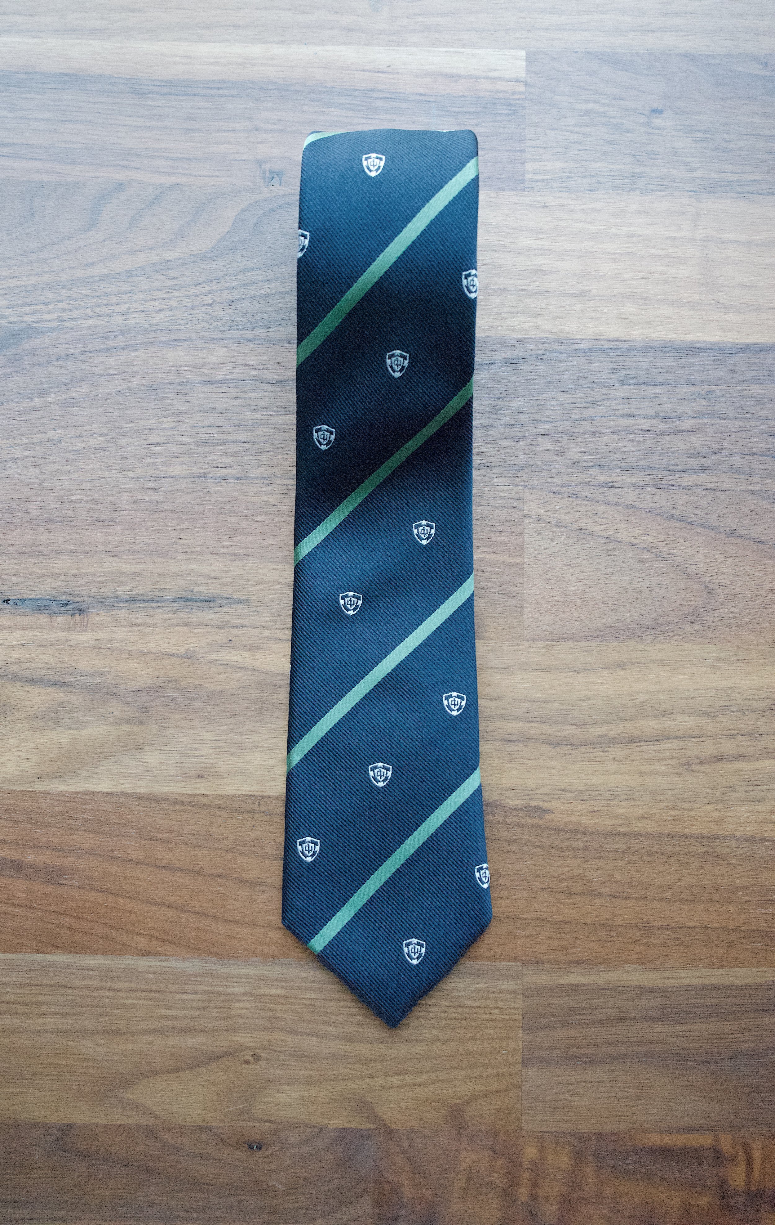 Navy necktie with green diagonal stripes and crest emblems laid flat on a wooden surface.
