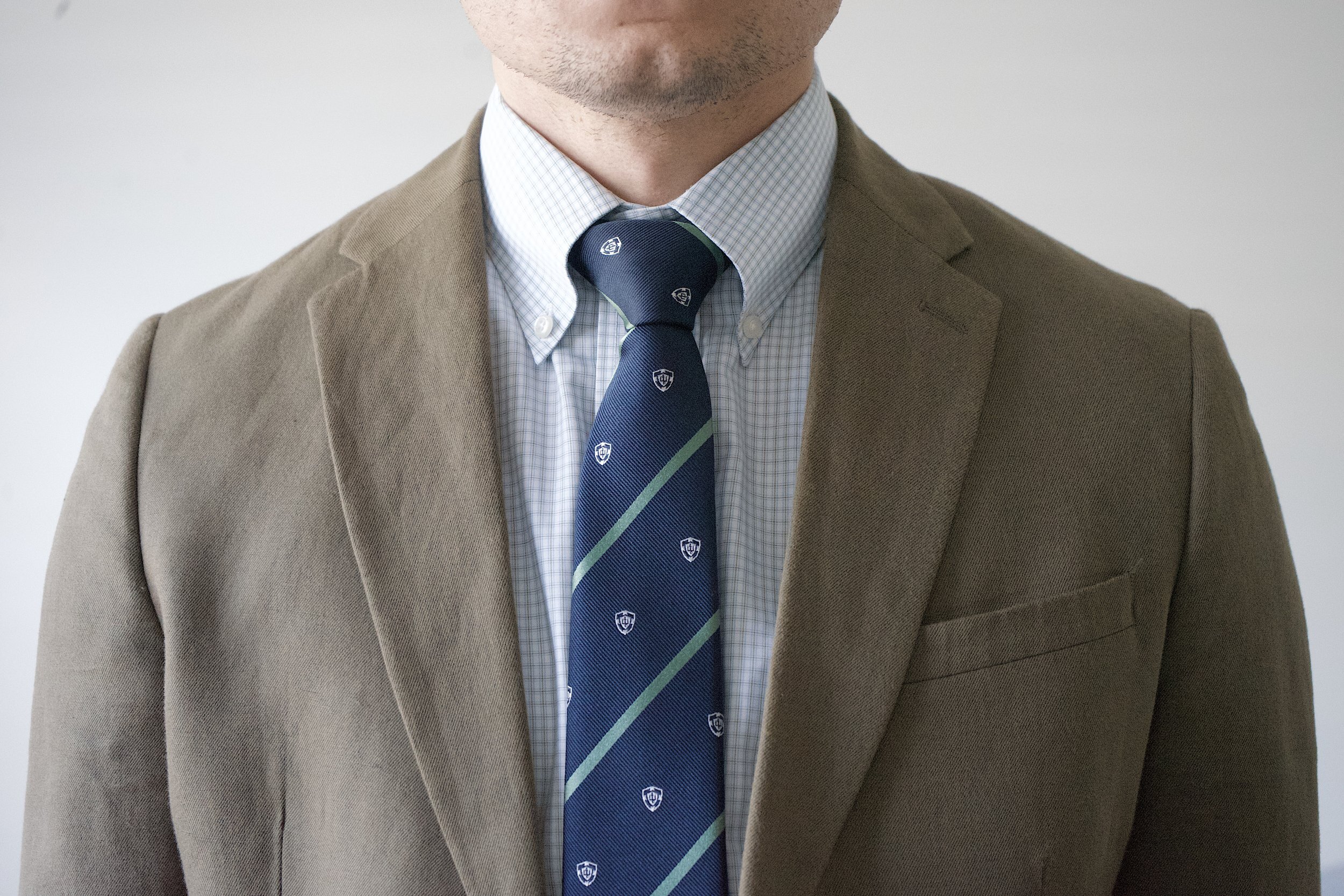 Navy necktie with green diagonal stripes and crest pattern, worn with a blazer and checkered dress shirt.