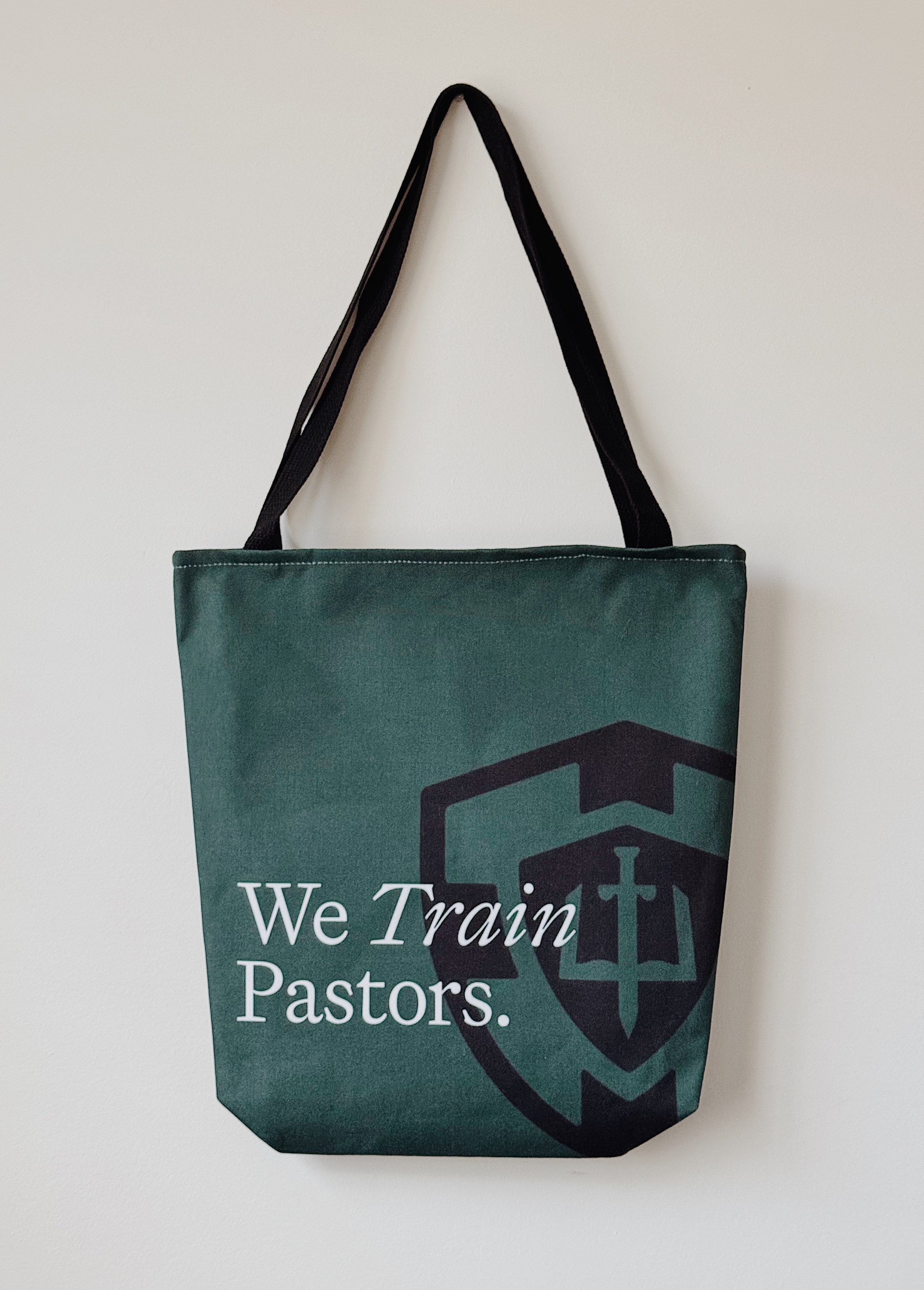 Green tote bag with large GPA-style crest graphic and the phrase ’We Train Pastors.’