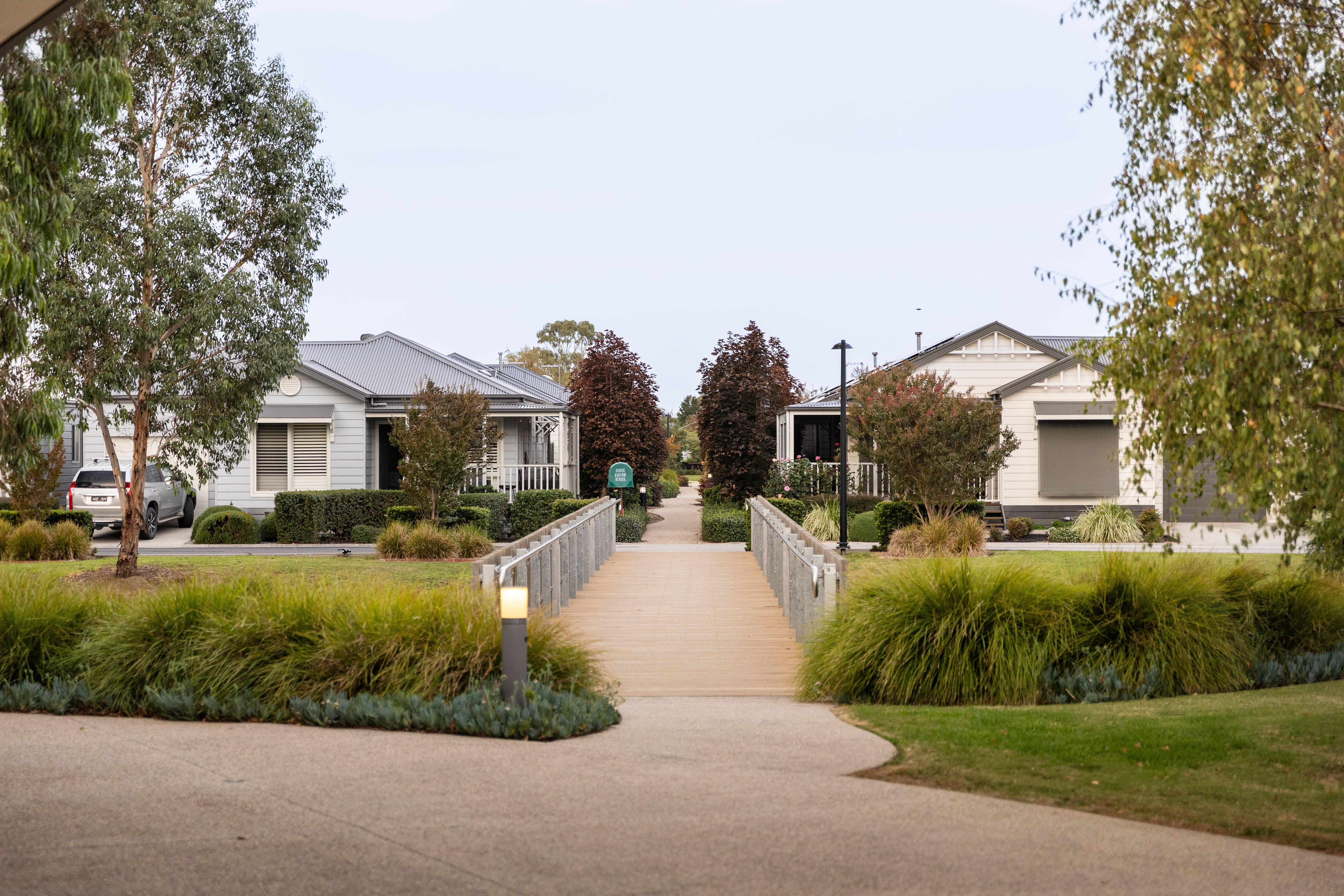 Pedestrian bridge and landscaped path between two modern homes surrounded by gardens and mature trees in a Lifestyle Communities neighbourhood.