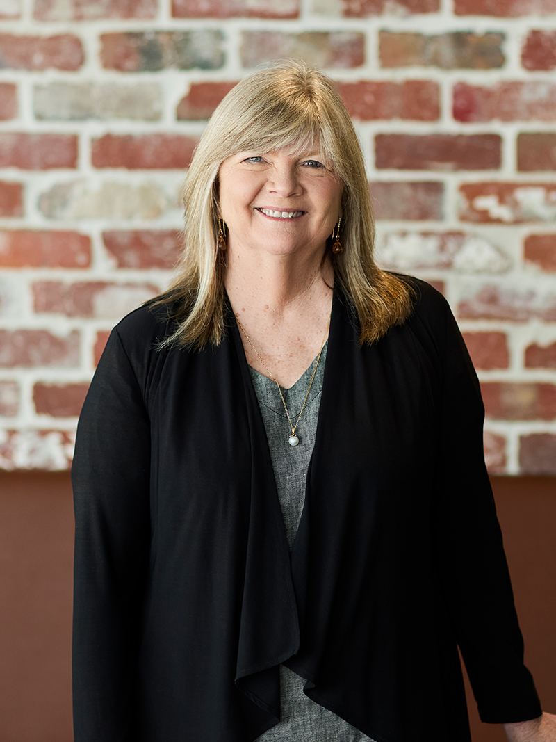 Professionally dressed individual standing indoors against an exposed brick wall.