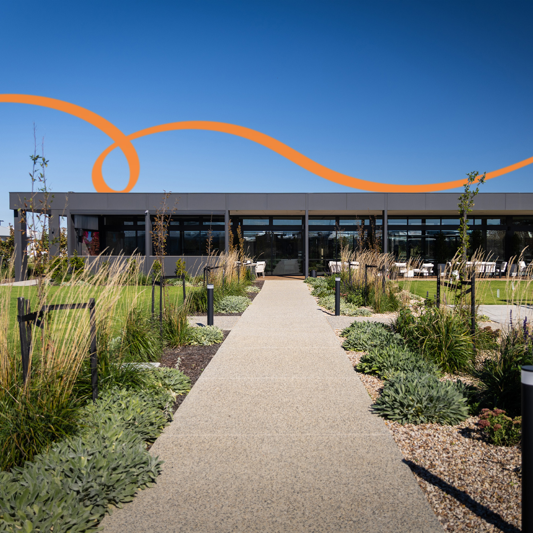 Pathway leading through landscaped gardens to the Ridgelea clubhouse, featuring outdoor seating, manicured lawns, and contemporary architecture under a clear blue sky.