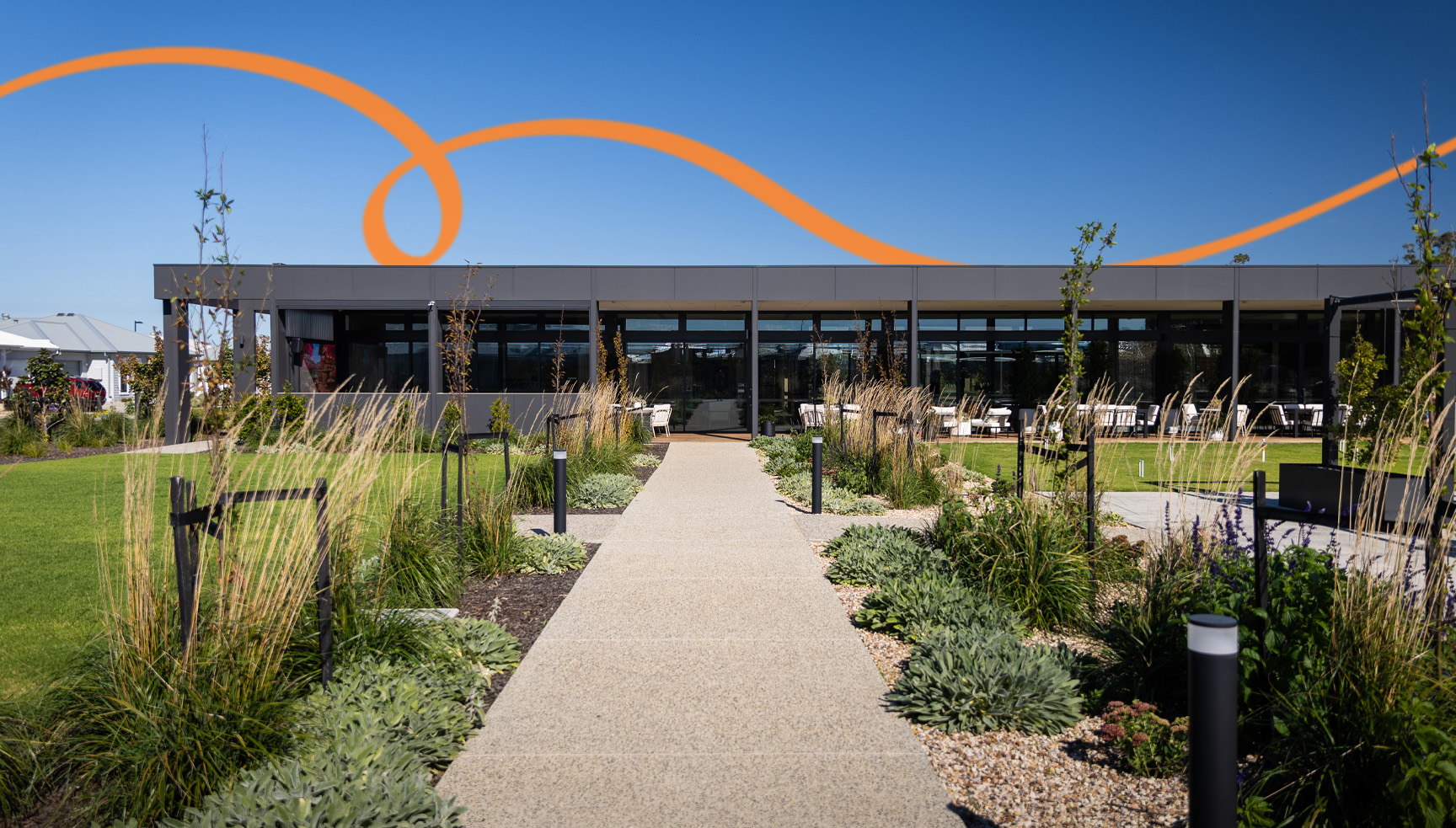 Pathway leading through landscaped gardens to the Ridgelea clubhouse, featuring outdoor seating, manicured lawns, and contemporary architecture under a clear blue sky.