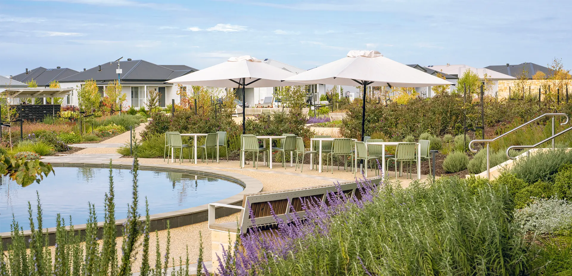 Landscaped community garden at Lifestyle Communities Woodlea featuring walking paths, flowering garden beds, outdoor seating areas with large umbrellas, and a central water feature surrounded by greenery.