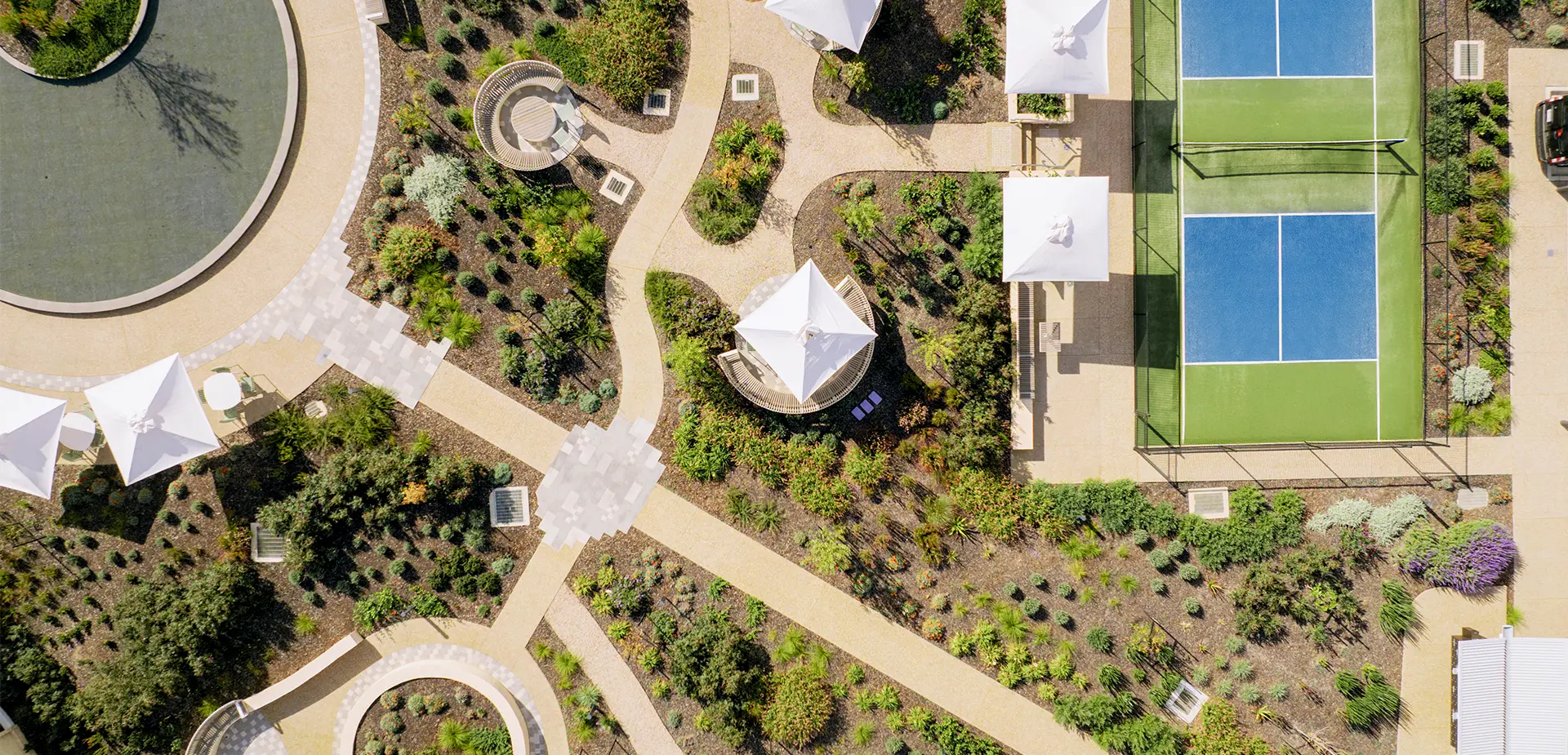 Aerial view of Lifestyle Communities Woodlea showing landscaped gardens, walking paths, shaded seating areas, a water feature, and adjacent tennis courts surrounded by greenery and communal outdoor spaces.