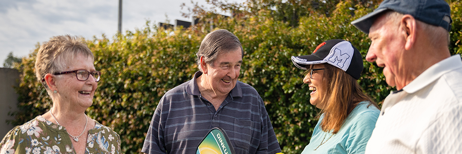 Four people standing outdoors in a Lifestyle Communities setting, chatting together in front of landscaped greenery and community homes.