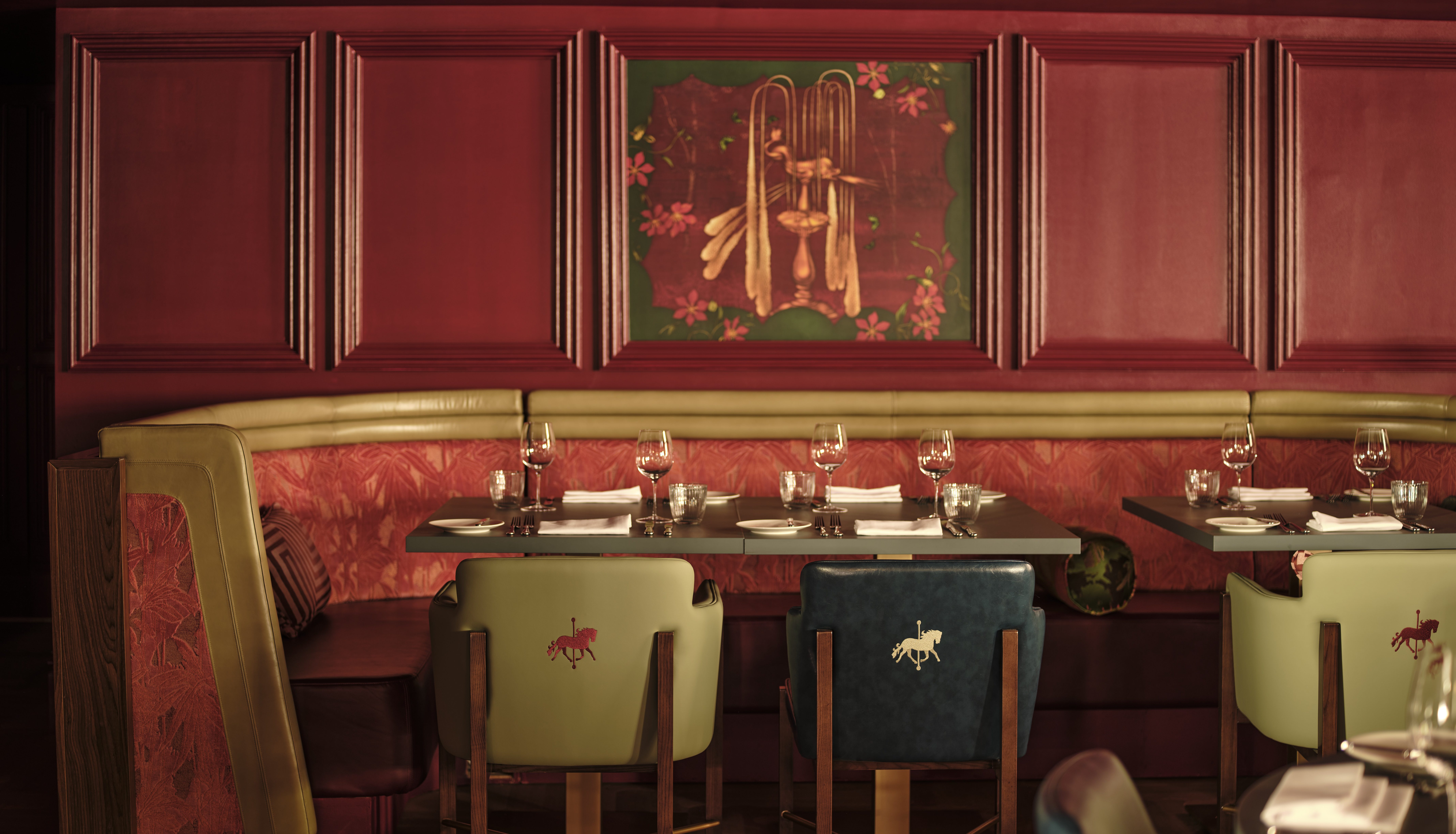 Restaurant booth with red panelled walls, gold trim banquette, set dining table, and mixed green and teal chairs with embroidered horse motifs.