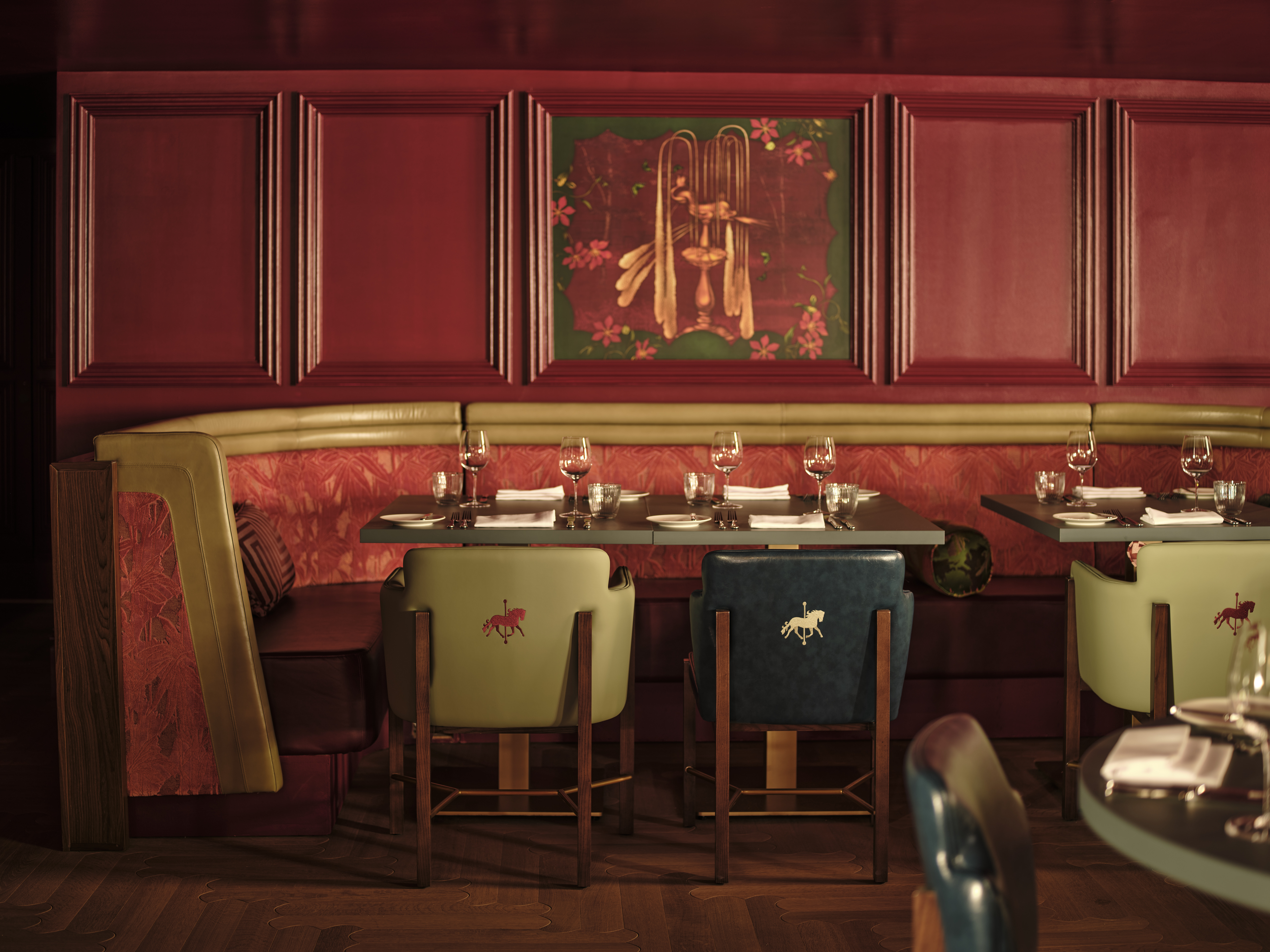 Restaurant dining area with red panelled walls, gold banquette seating, green and teal chairs with embroidered horses, set tables and a decorative wall painting.