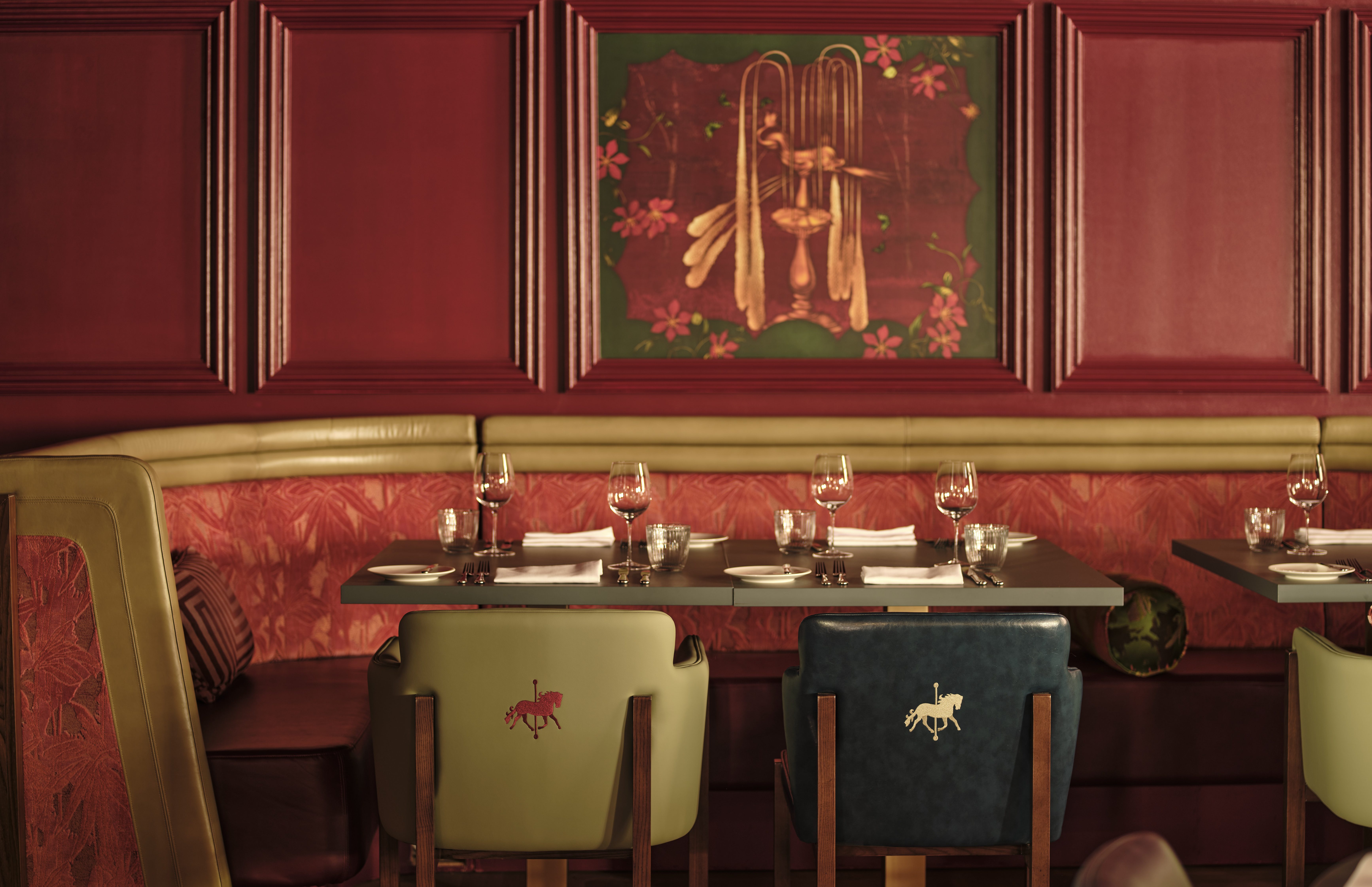 Restaurant dining area with red panelled walls, gold banquette seating, set tables with wine glasses, and chairs embroidered with a horse motif.