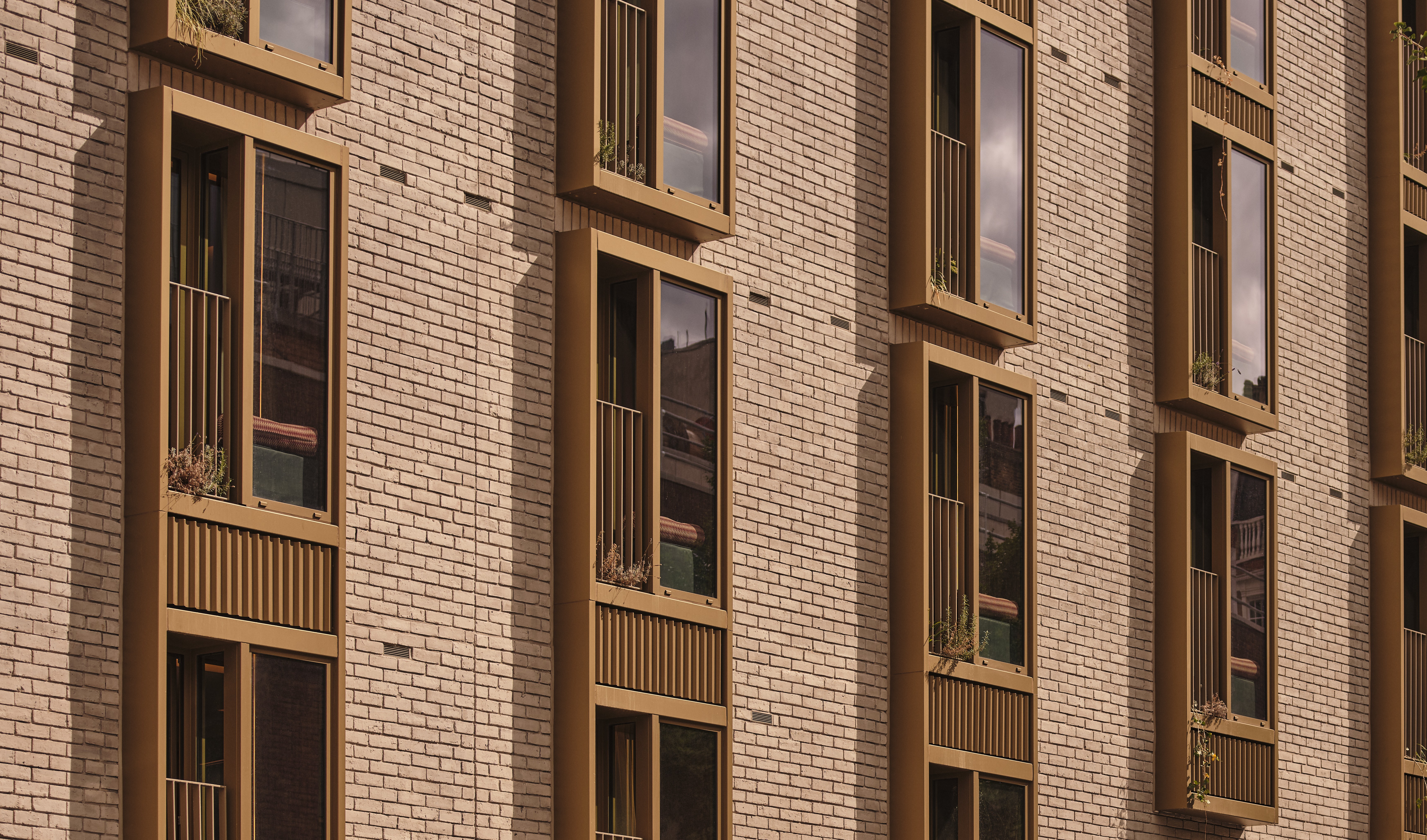 Close-up of a residential building facade with light beige brick, warm brown-framed windows, and small Juliet balconies with vertical metal railings.