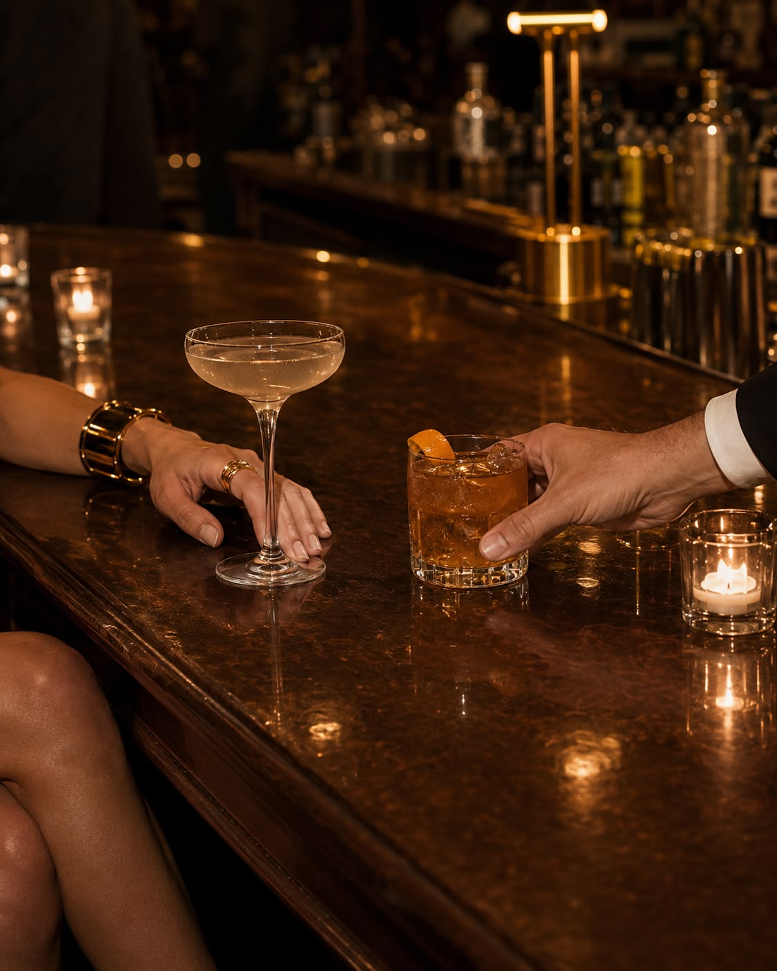 Two hands reach for cocktails on a dark wooden bar. A coupe glass and an amber whisky drink sit beside a lit candle, bottles visible behind.