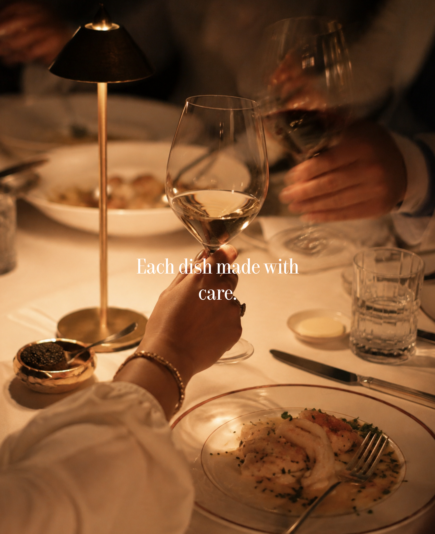 Hand holding a white wine glass at a restaurant table with a small brass lamp, caviar dish, and a plated starter.