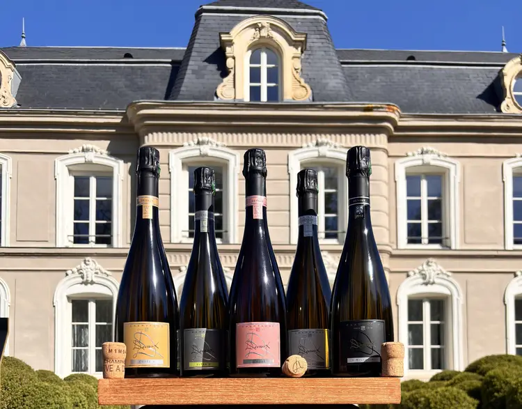 Five champagne bottles on a wooden stand in front of a cream stone French château under a clear blue sky.