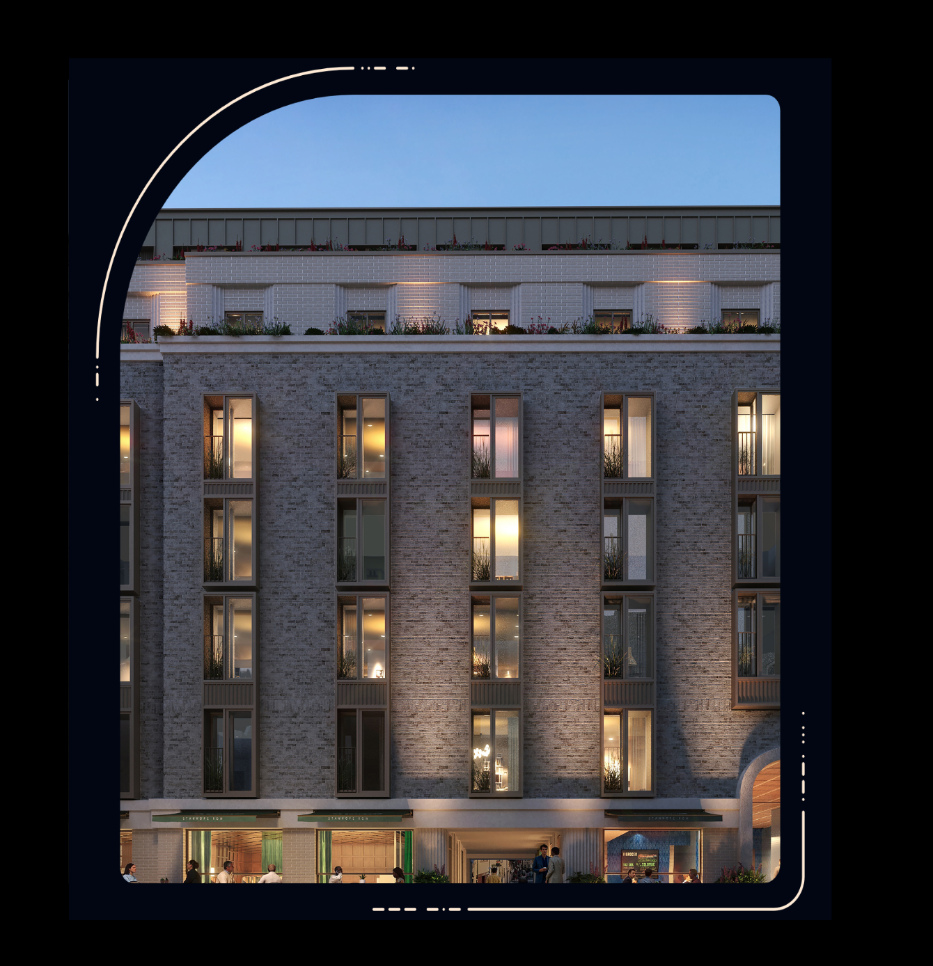 Multi-storey brick building at dusk with illuminated windows, rooftop terrace with greenery, and ground-floor retail spaces with people outside.