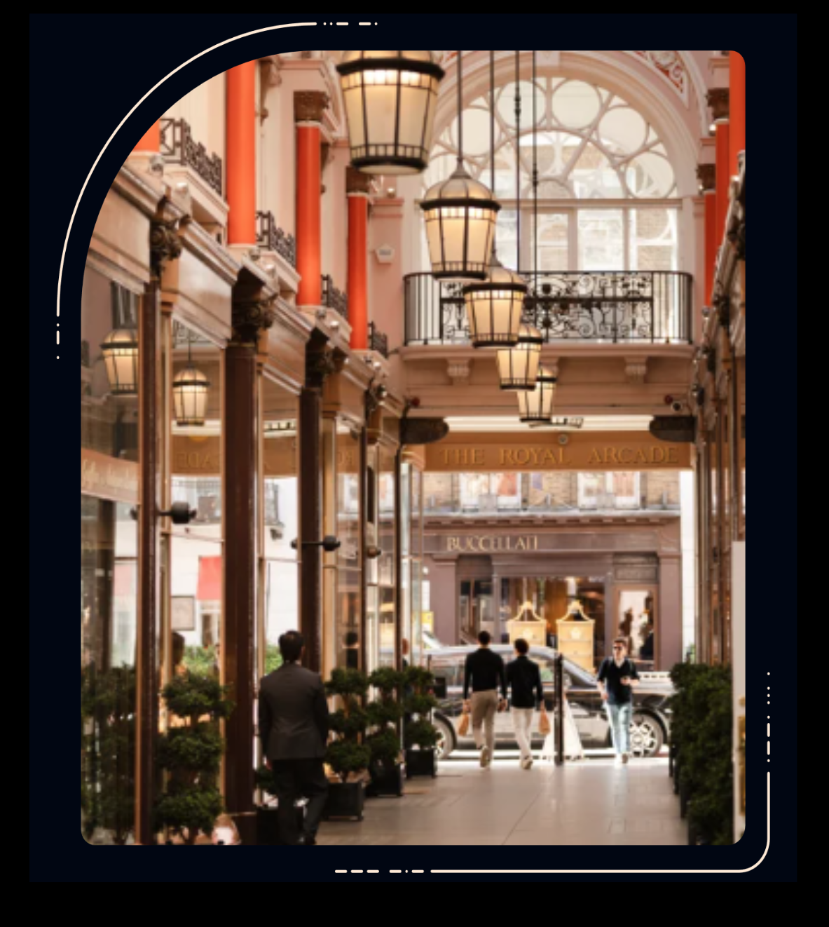 Interior of an elegant shopping arcade with hanging lanterns, red columns, ornate architecture, and people walking towards the entrance.