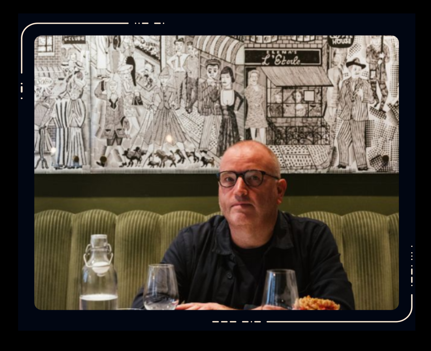 Man wearing glasses and dark shirt seated at a restaurant table with glasses and a carafe, green banquette seating and black-and-white mural behind him.