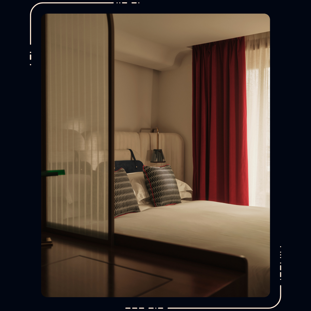 Hotel room with double bed, patterned cushions, cream headboard, red curtains by a window, viewed through a glass partition.