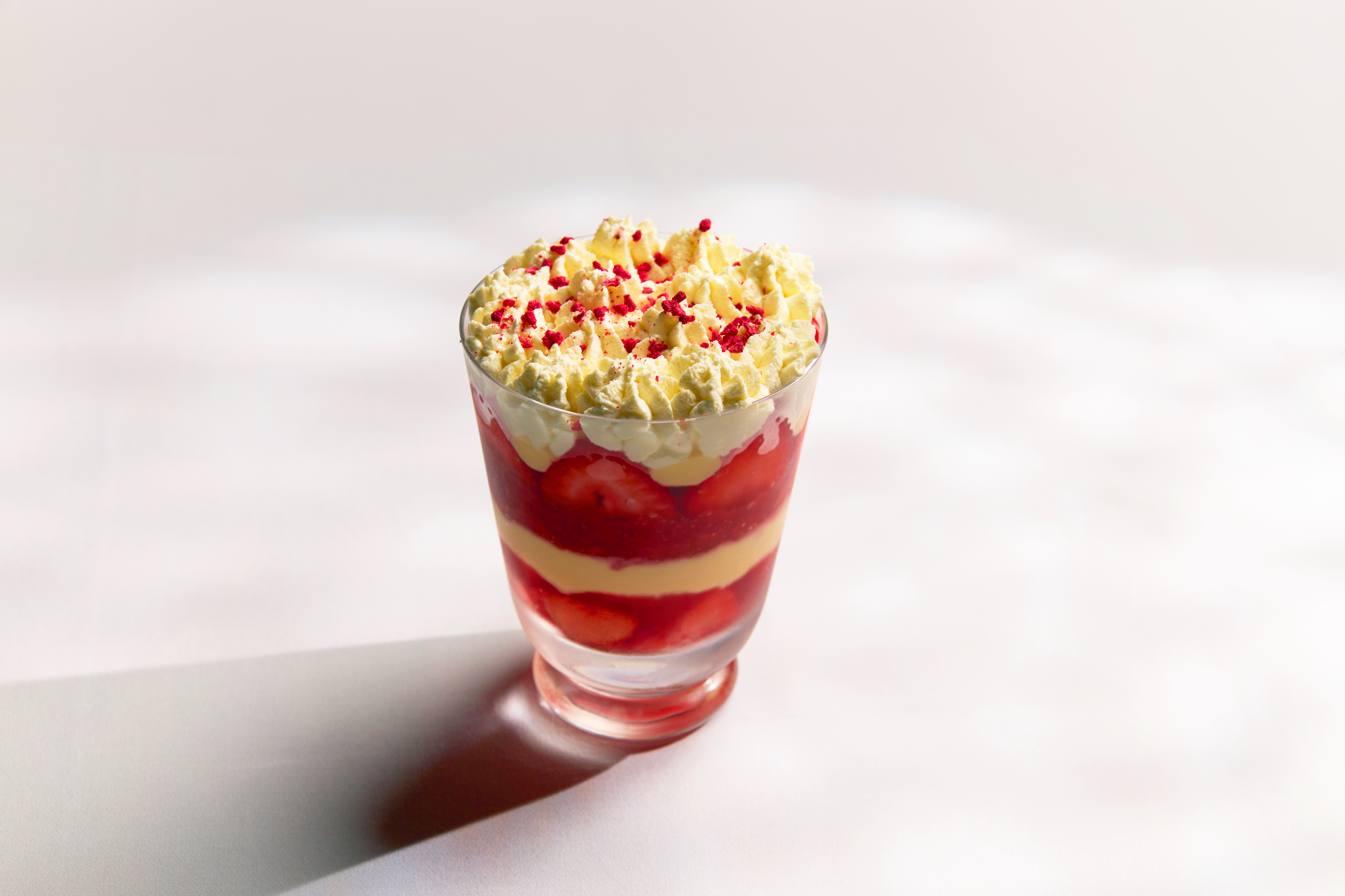 Layered dessert in a glass with red jelly, custard, and whipped cream topped with red sugar sprinkles, on a white background.