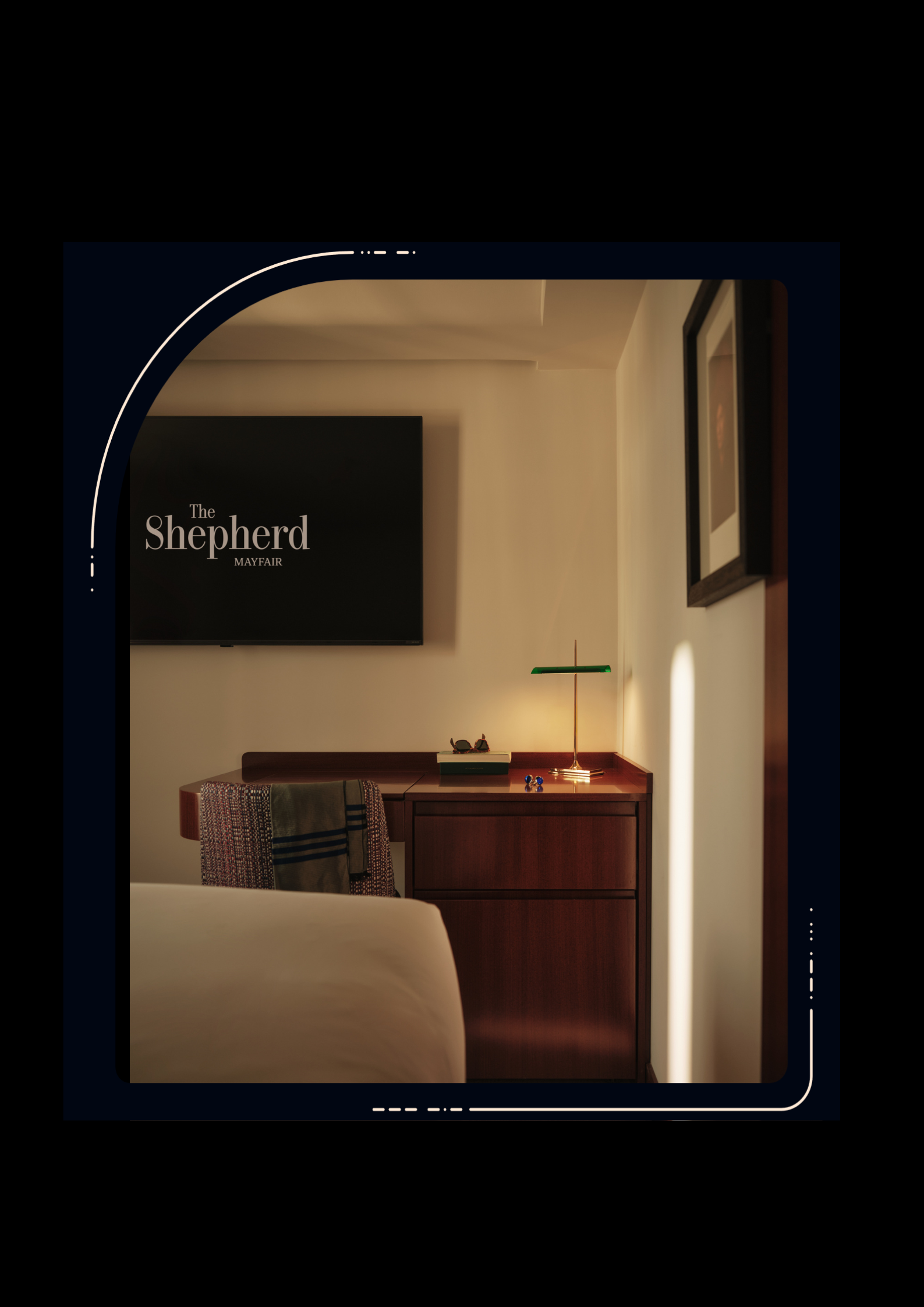Hotel room at The Shepherd Mayfair showing a wooden bedside cabinet, small green desk lamp, wall-mounted TV, framed artwork and a chair with a blanket.