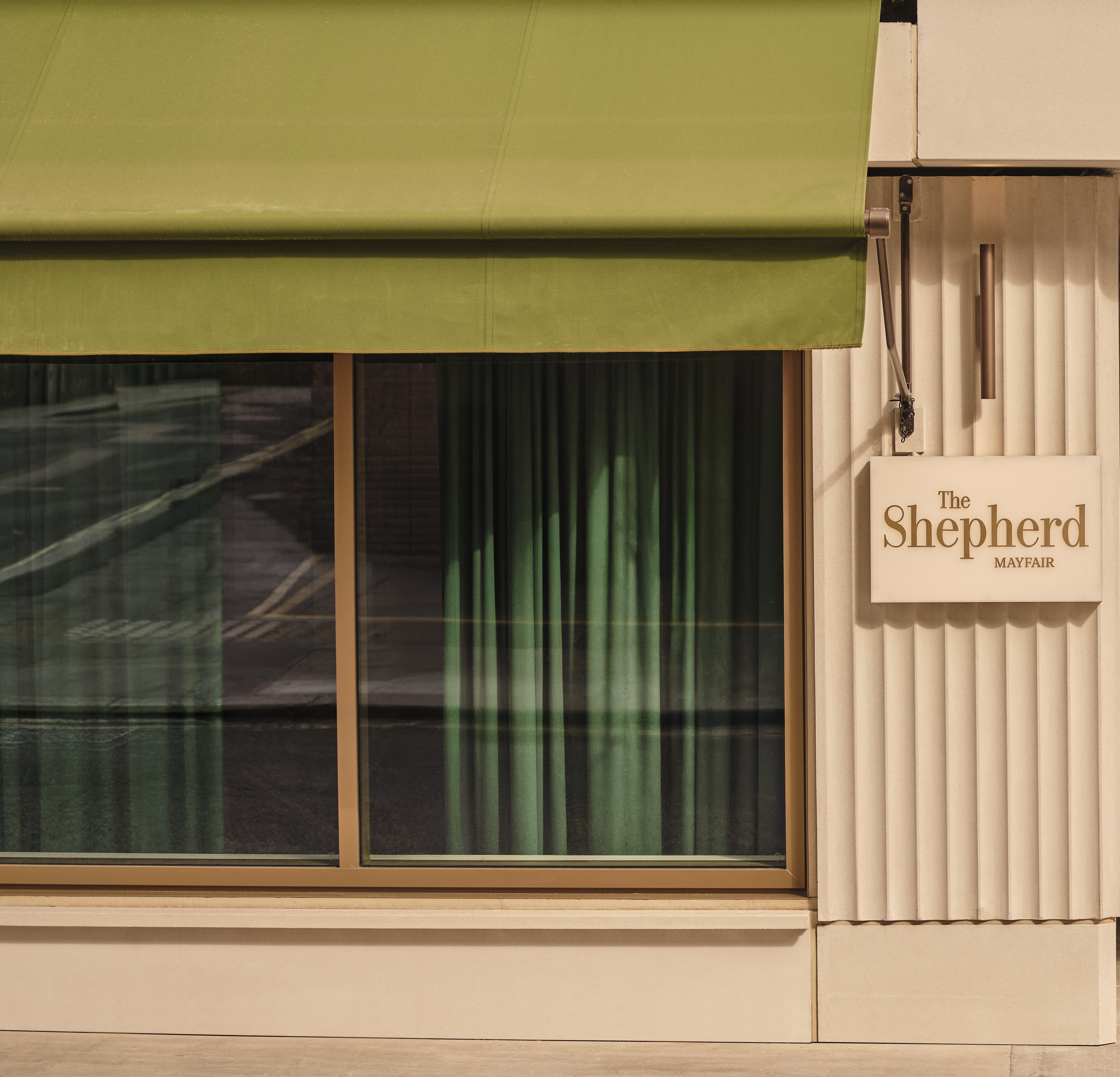 Exterior of The Shepherd Mayfair, showing a green awning, large window with green curtains, and a wall-mounted sign.