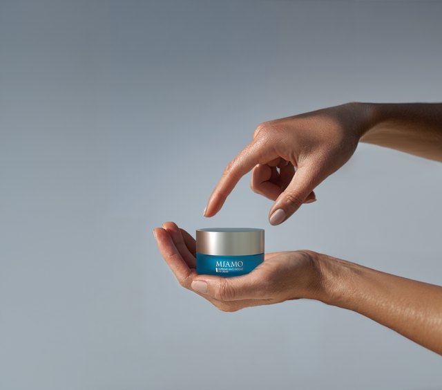 Two hands holding and pointing to a small teal and silver Miamo branded skincare cream jar against a grey background.