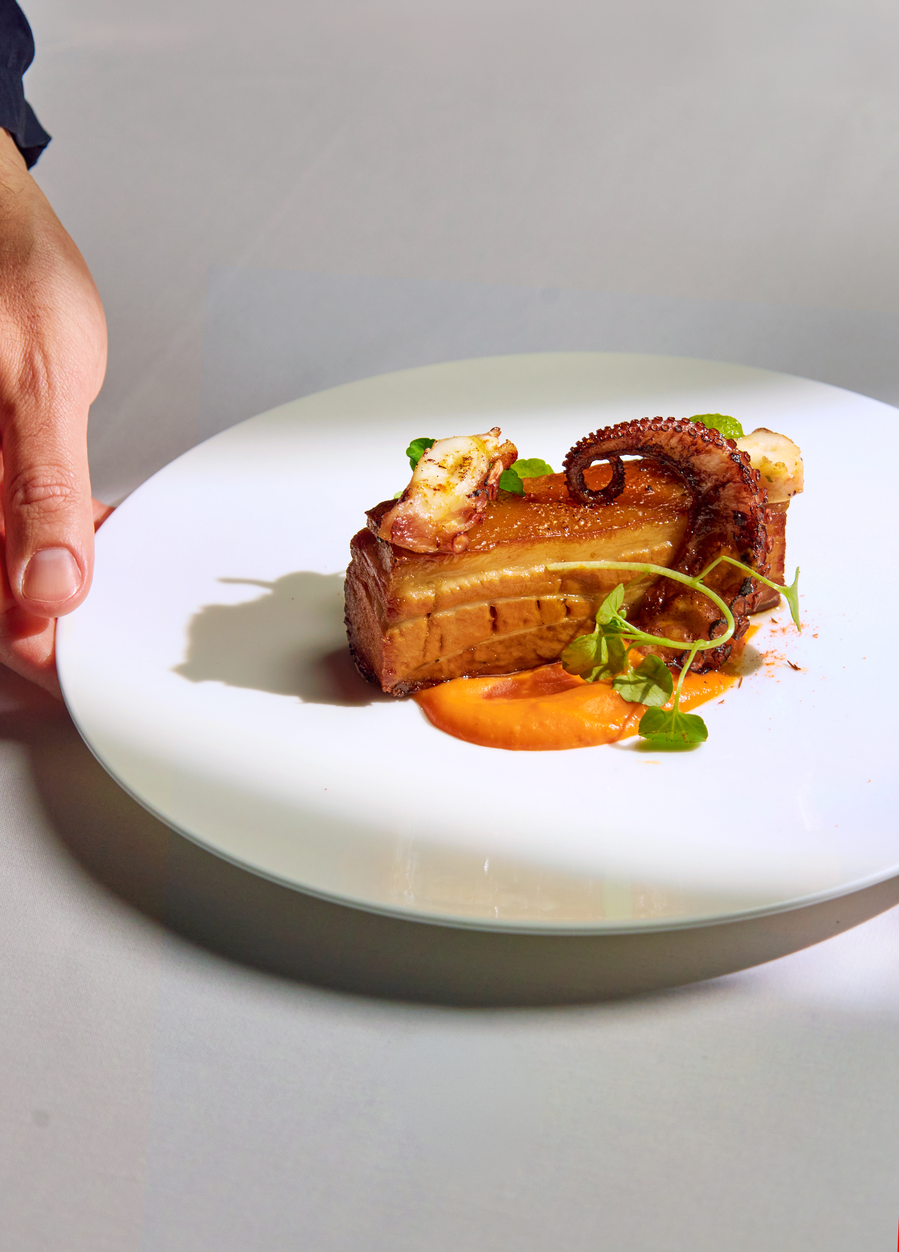 Crispy pork belly topped with grilled octopus tentacle, orange purée, and micro herbs on a white plate, held by a hand.