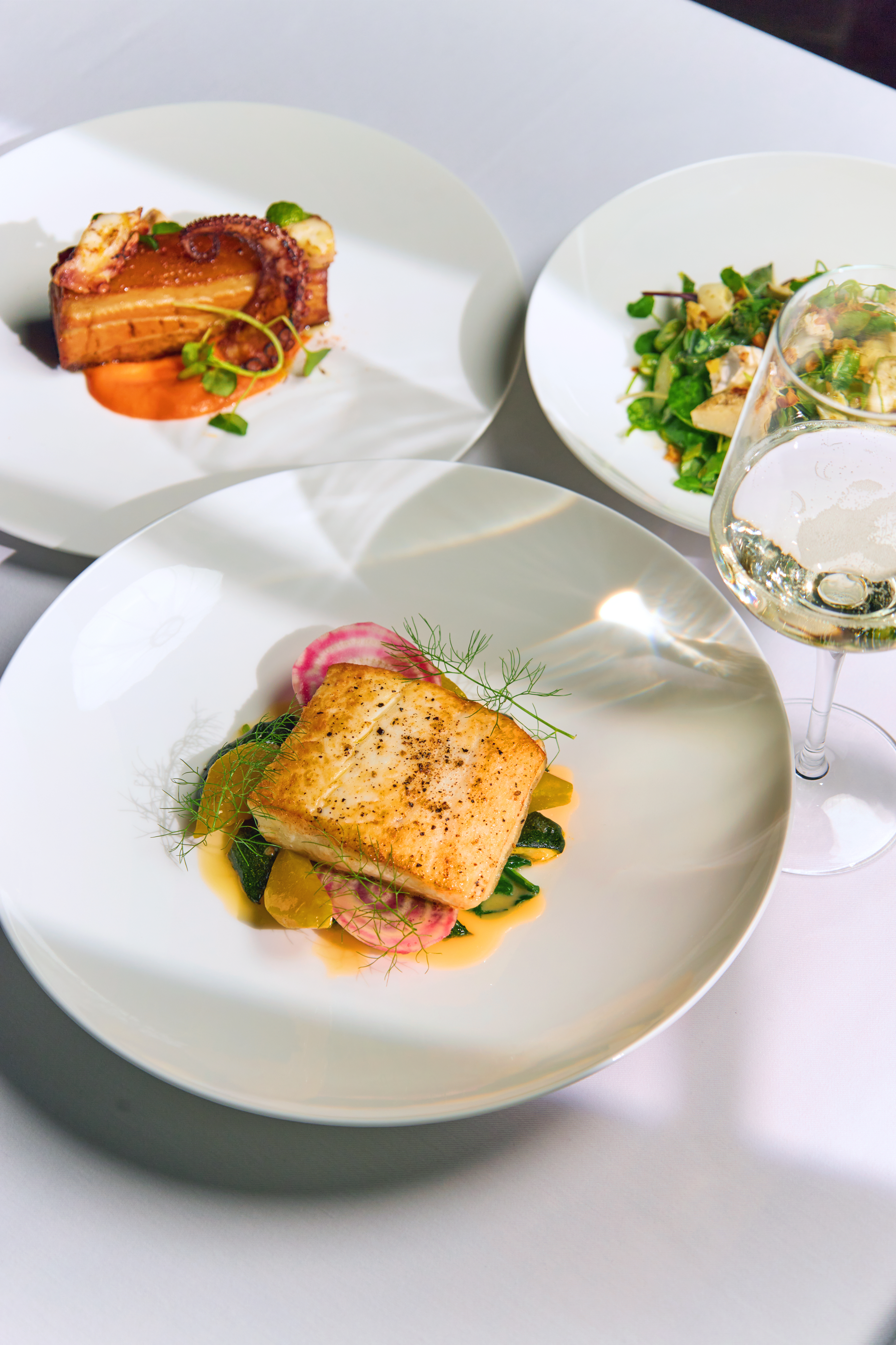 Three plated restaurant dishes on white table: seared fish with radish and greens, pork belly with octopus, and a salad. White wine glass alongside.