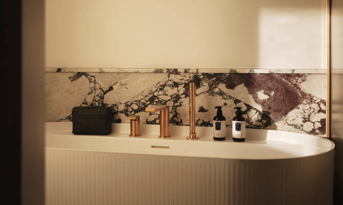 Freestanding bath with ribbed panel, rose gold taps, marble wall with purple veining, black toiletry bag and two white pump bottles.