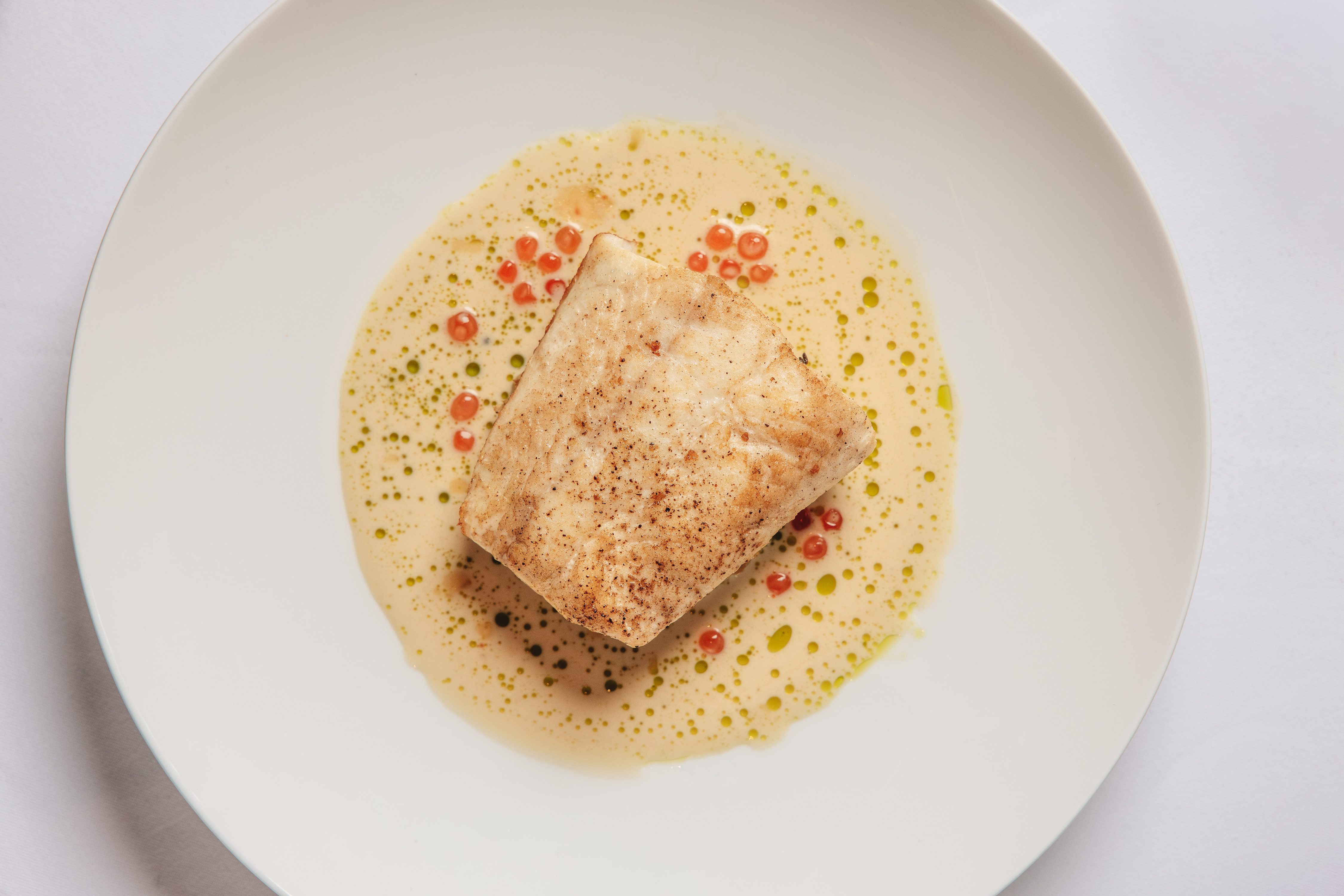 Seared fish fillet in a cream sauce with red caviar and green oil drops, served in a wide white bowl.