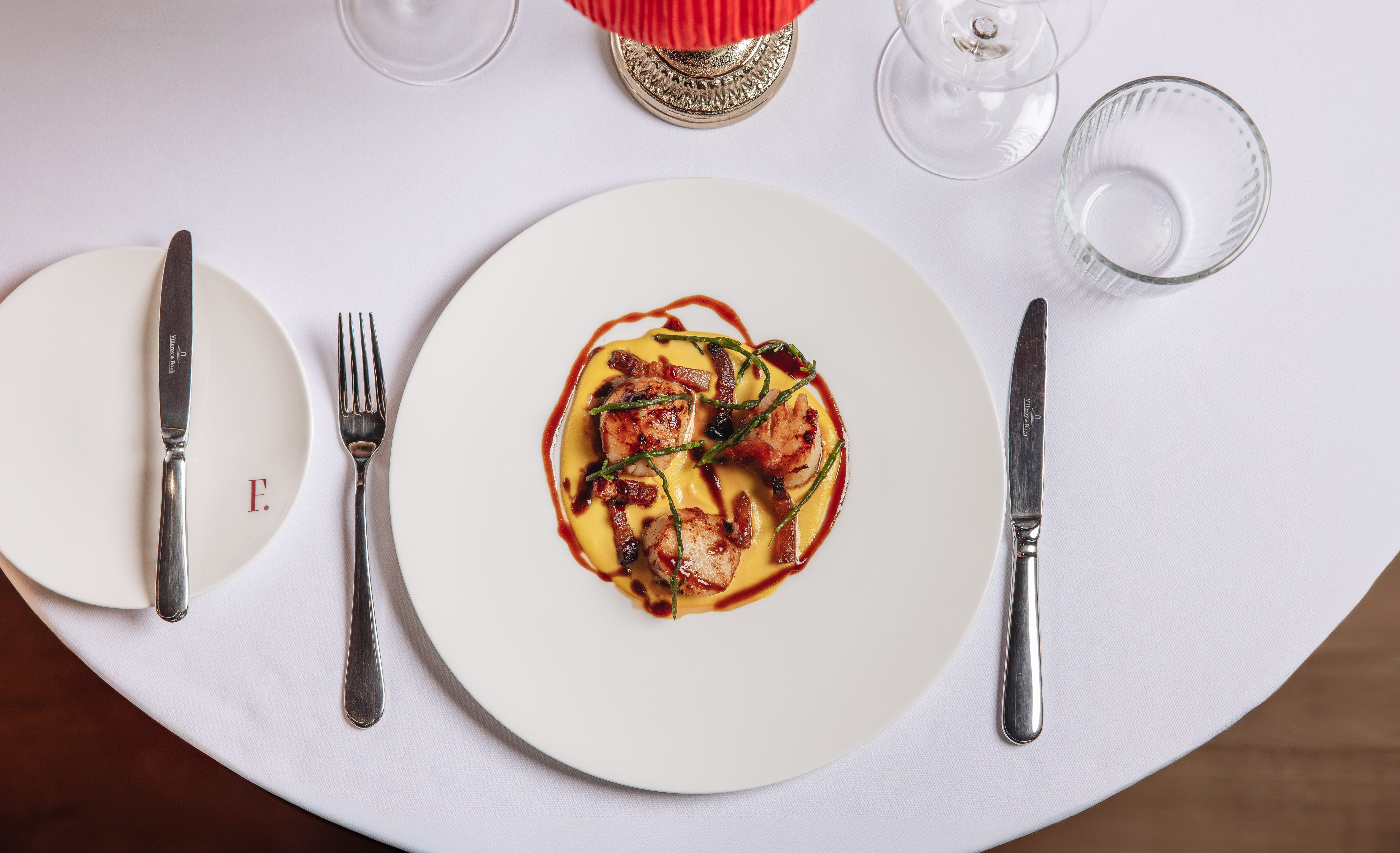 Overhead view of a plated restaurant dish with meat and red sauce on a white tablecloth, flanked by cutlery and glassware.