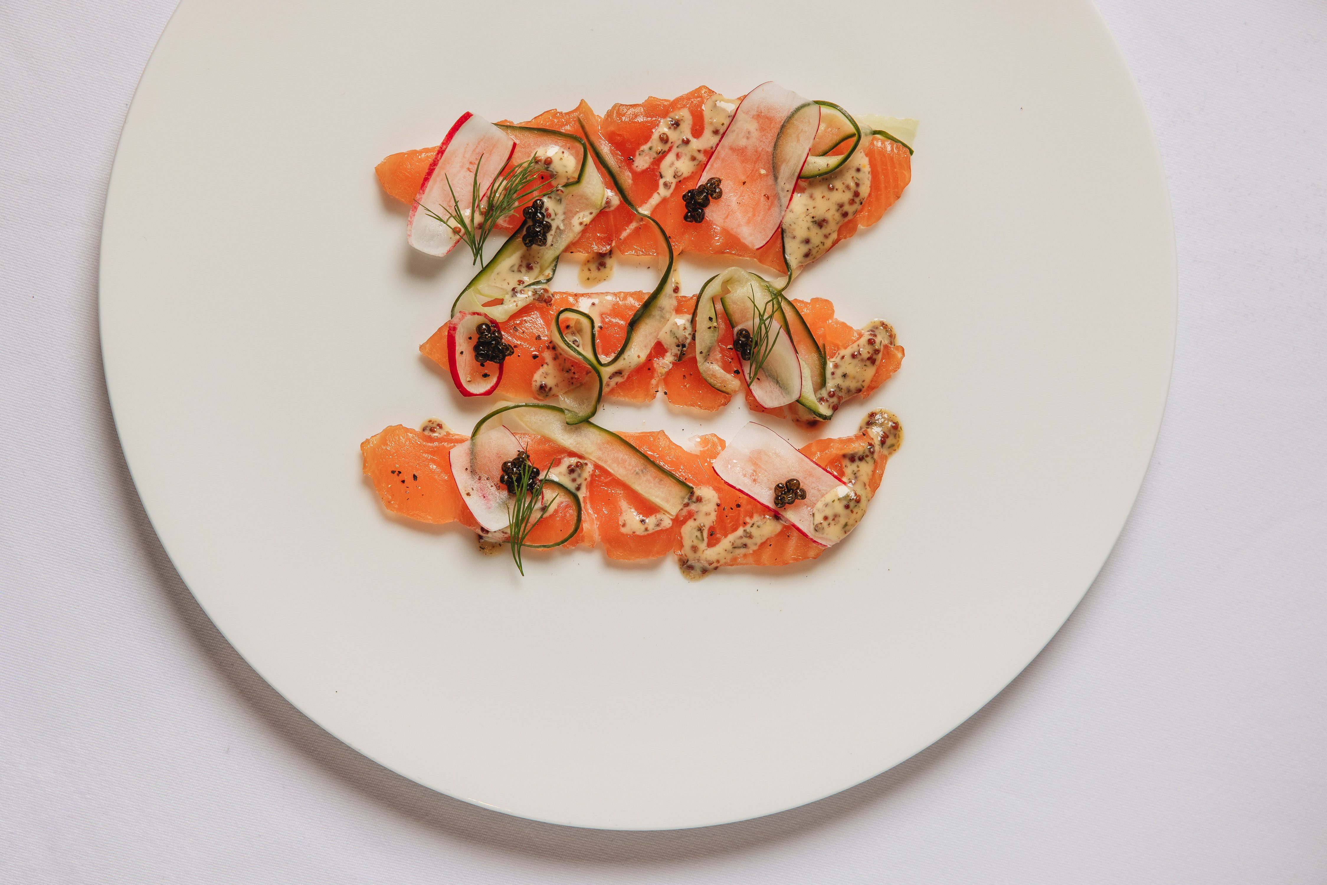 Overhead view of cured salmon slices on a white plate, garnished with cucumber ribbons, radish, dill, caviar, and a mustard sauce.