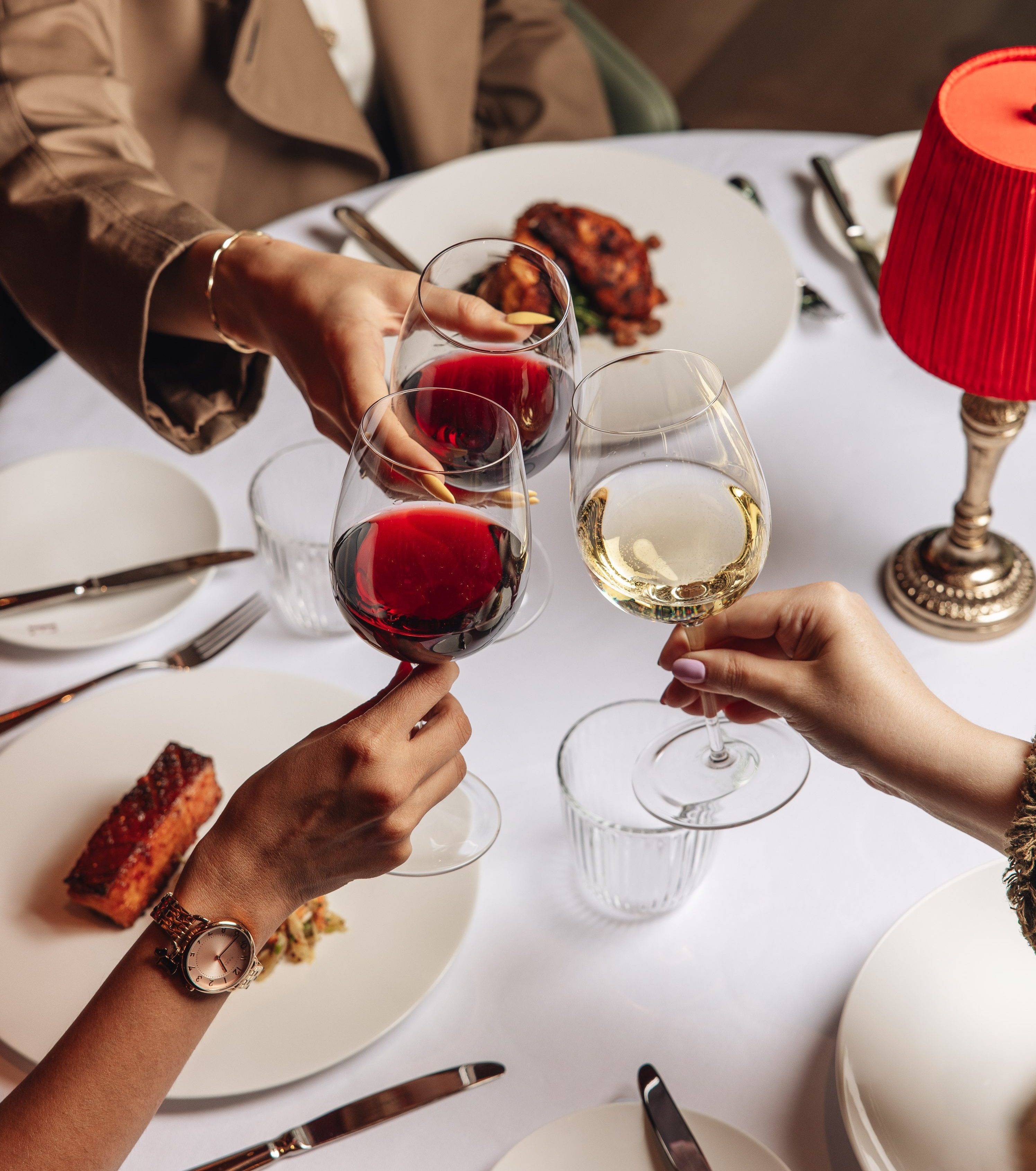 People clinking glasses of red and white wine over a white restaurant table set with cutlery, plates of food, and a red table lamp.