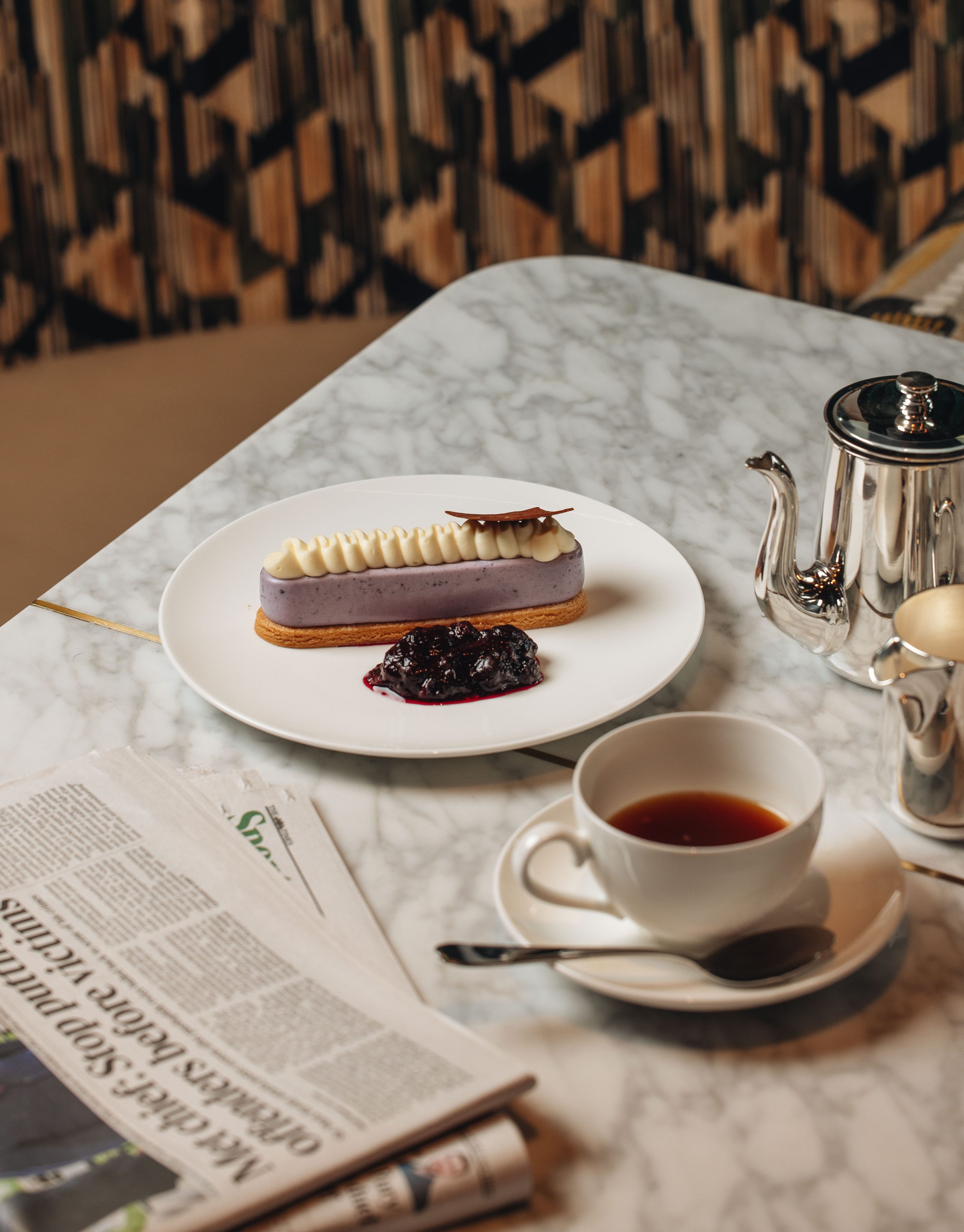 Marble table with a purple éclair on a white plate, cup of tea, silver teapot, and a folded newspaper. Geometric patterned wall behind.