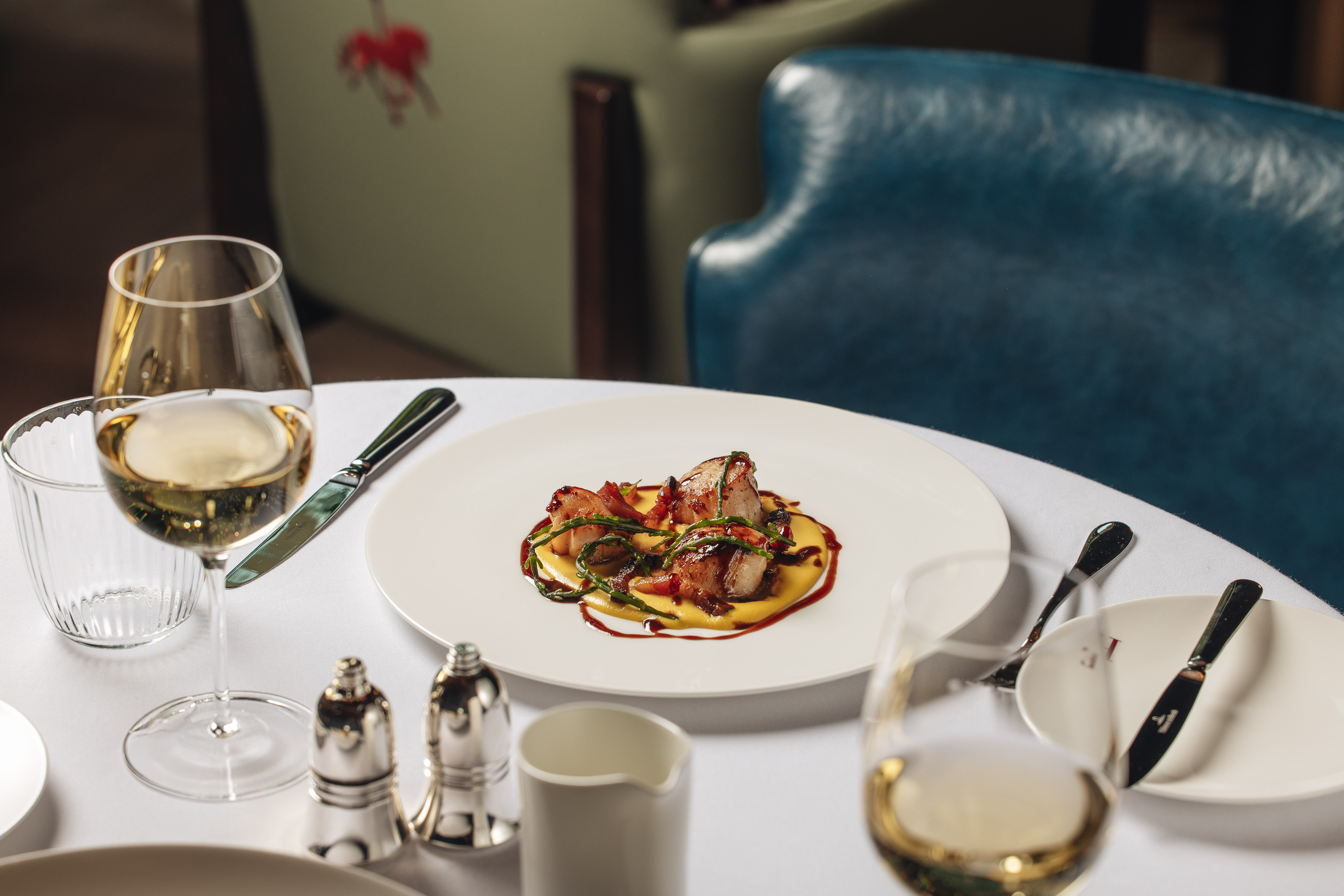 A plated restaurant dish with meat and vegetables on a white round table, with white wine glasses, cutlery, and condiments nearby.