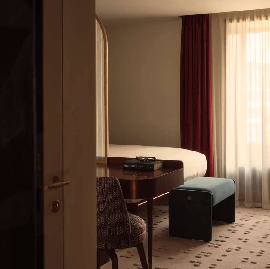 Hotel room with a wooden desk, woven chair, blue ottoman, white bed, patterned carpet, and burgundy curtains against sheer drapes.