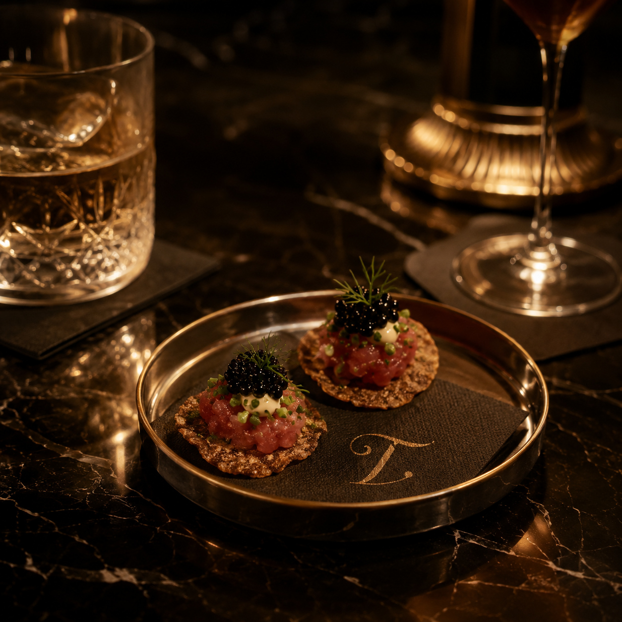 Two tartare canapés topped with caviar and dill on a dark gold-rimmed tray with engraved T, beside crystal glassware on marble.