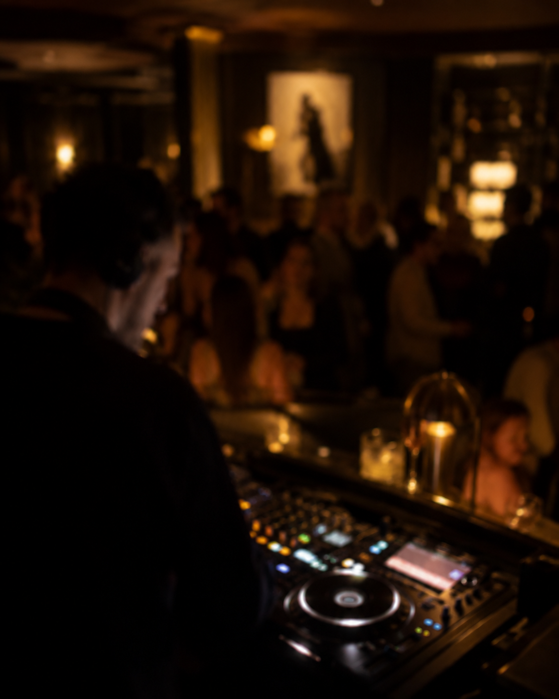 DJ at decks in a dimly lit venue, facing a crowd. Warm amber lighting, blurred background with people gathered.