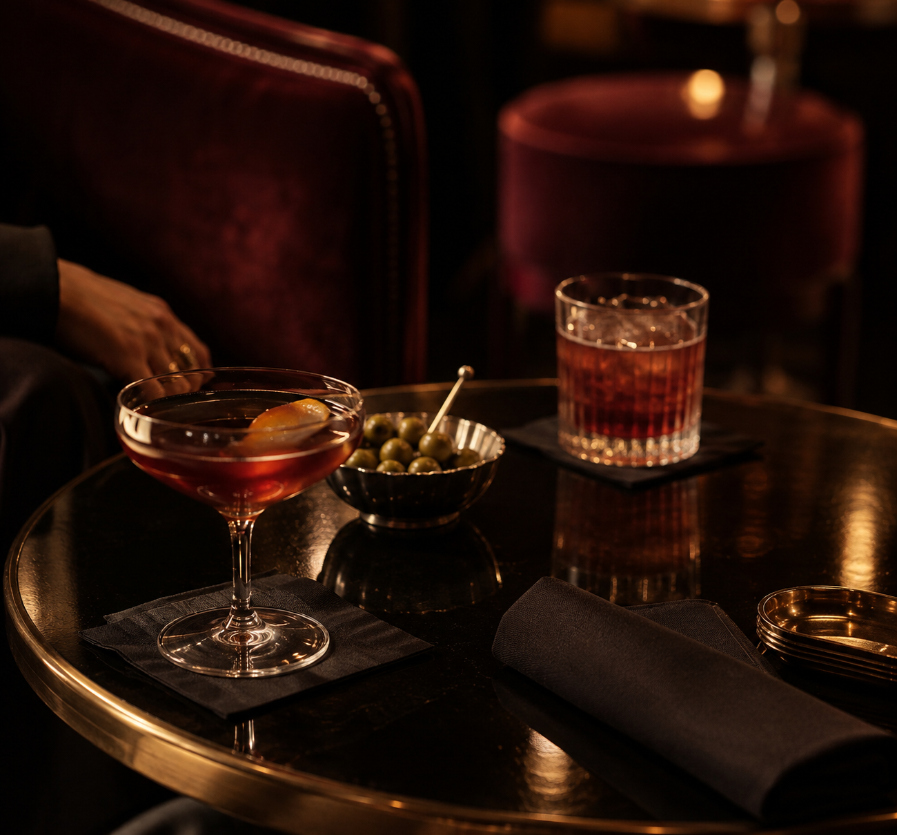 Two cocktails on a dark bar table: a coupe glass with amber drink and a rocks glass with ice, beside a bowl of olives.