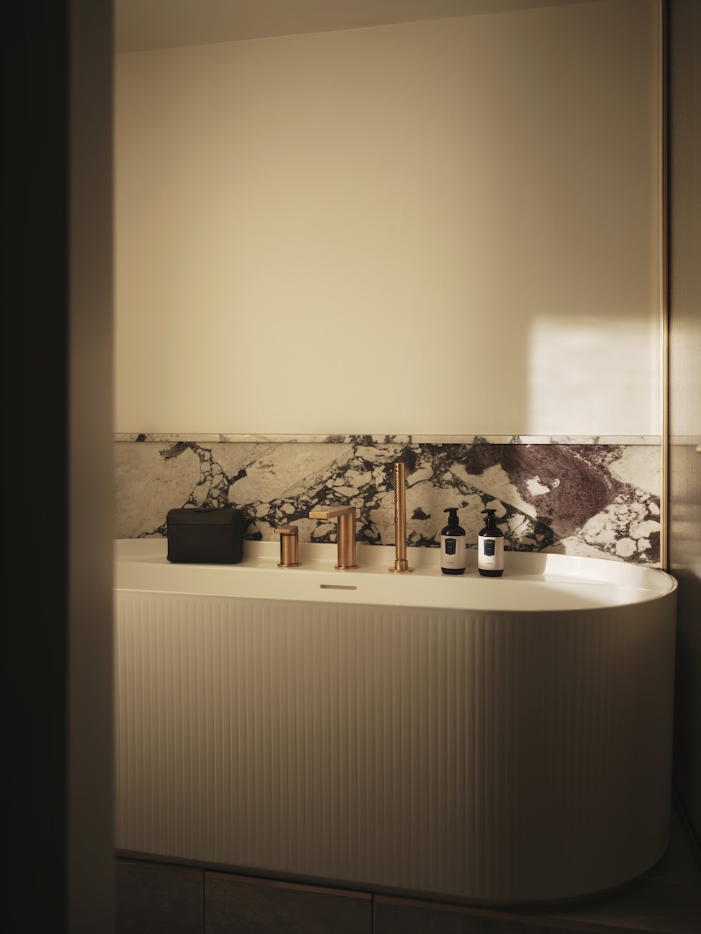 Modern bathroom with a fluted white bathtub, brass fixtures, and a marble backsplash, lit warmly.