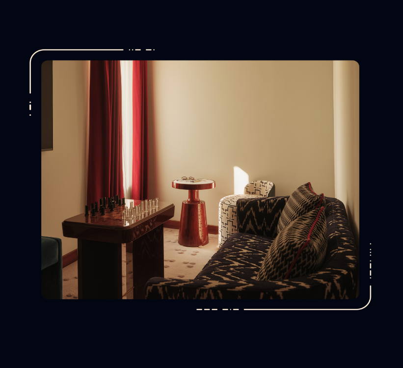 Hotel room seating area with patterned dark sofa, chess set on wooden table, red side table, armchair, and red curtains.