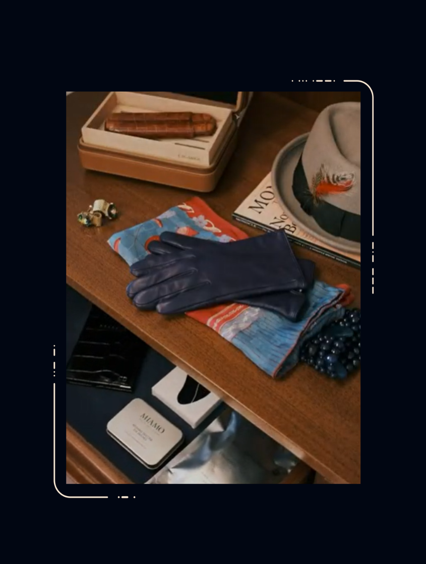 Navy leather gloves resting on a colourful scarf on a wooden dresser, with a fedora hat, cigar box, cufflinks and small tin nearby.