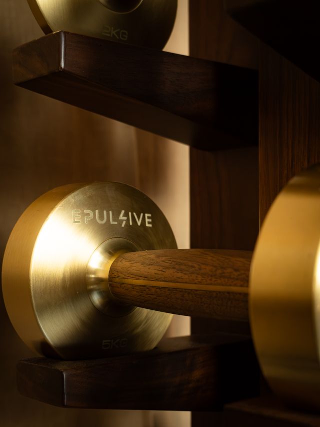 Close-up of Epulsive-branded dumbbell with brushed gold ends and dark wood handle, stored on a dark wooden rack.